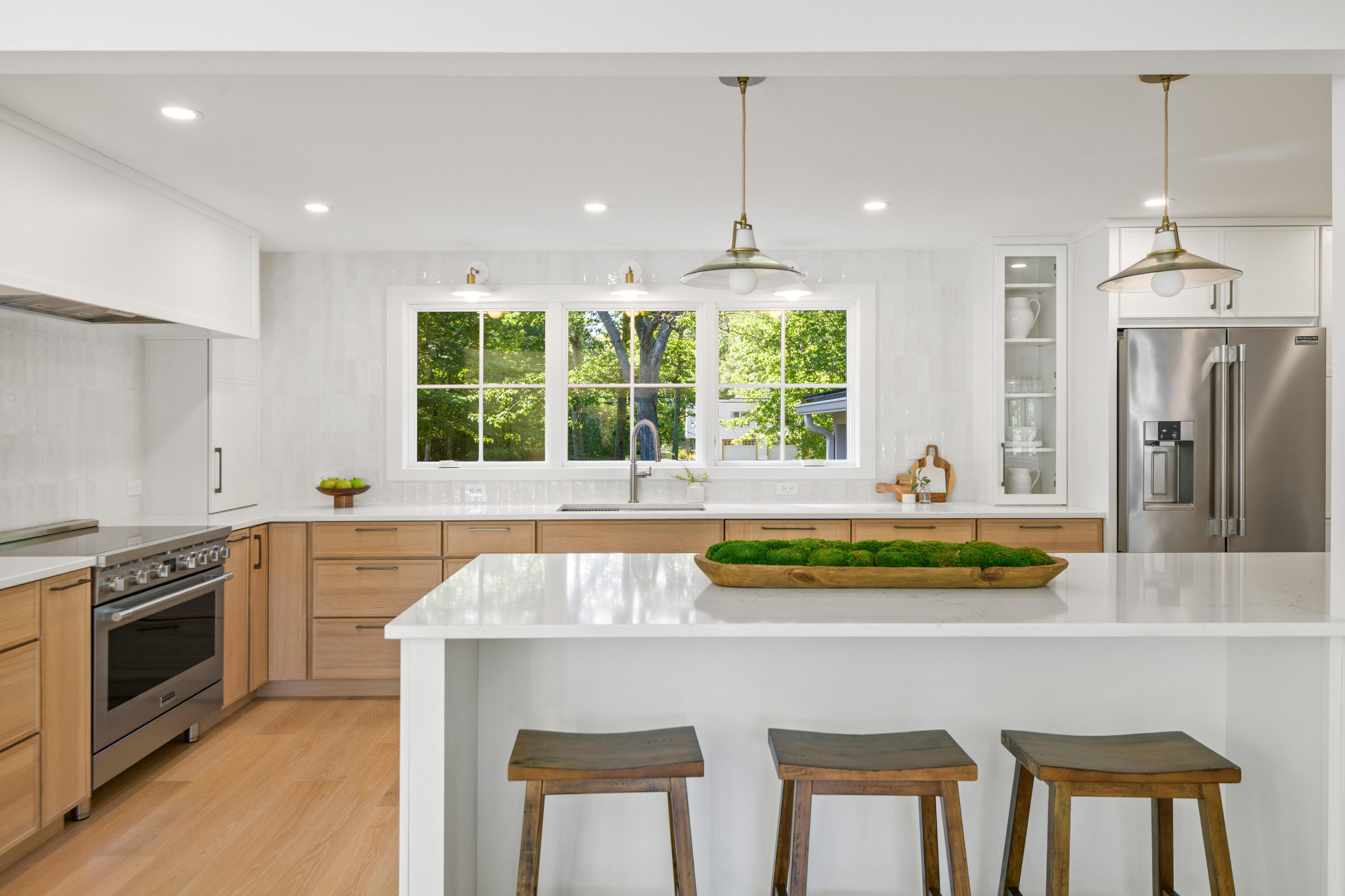 Bright kitchen with a large white island, three wooden stools, light wood cabinetry, and a window overlooking trees.