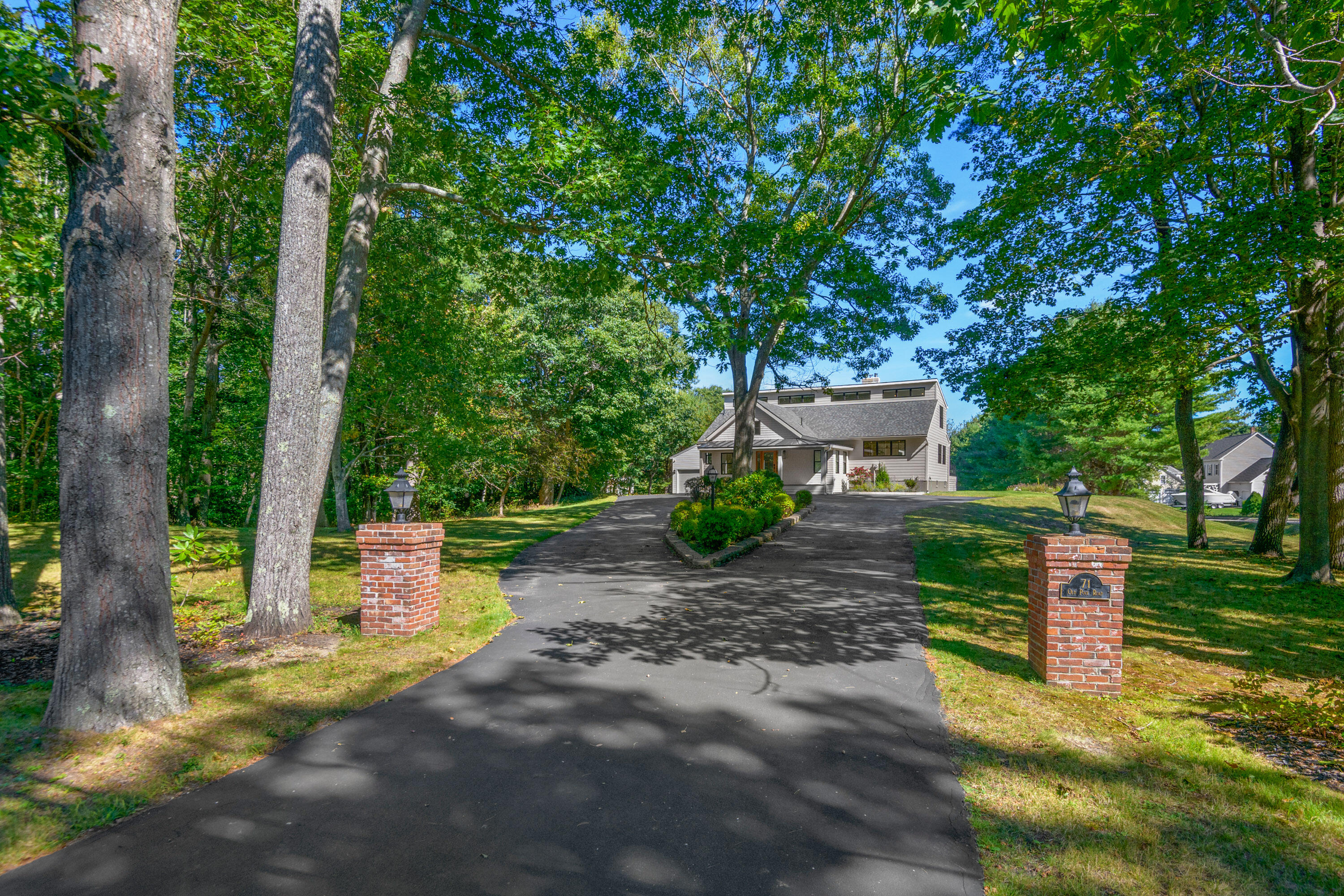 Gray house at the end of a tree-lined driveway, framed by brick entrance pillars and mature trees.
