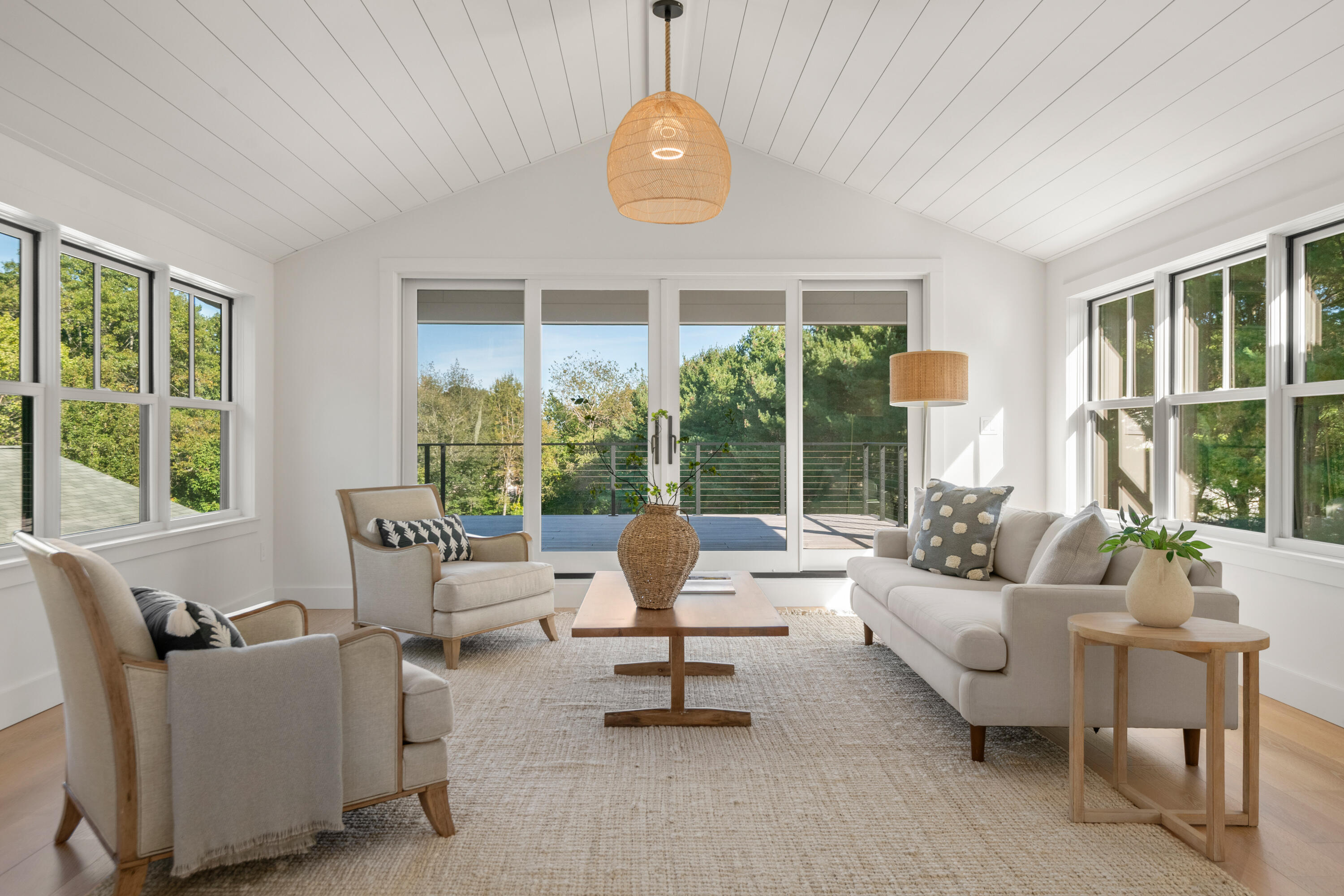Staged living room with a vaulted ceiling, large windows, and sliding doors to a wooded deck.