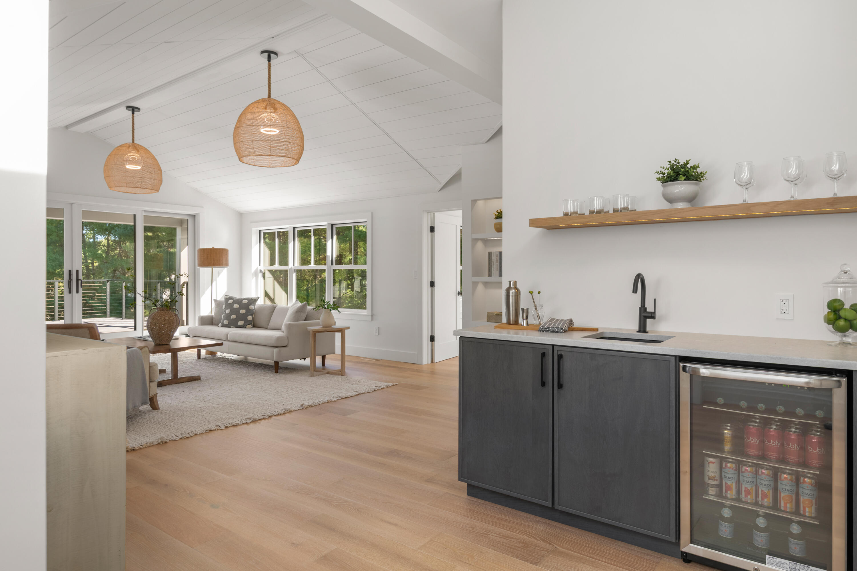 Open living room and wet bar with vaulted ceiling, woven pendants, dark cabinetry, and doors to a tree-lined deck.