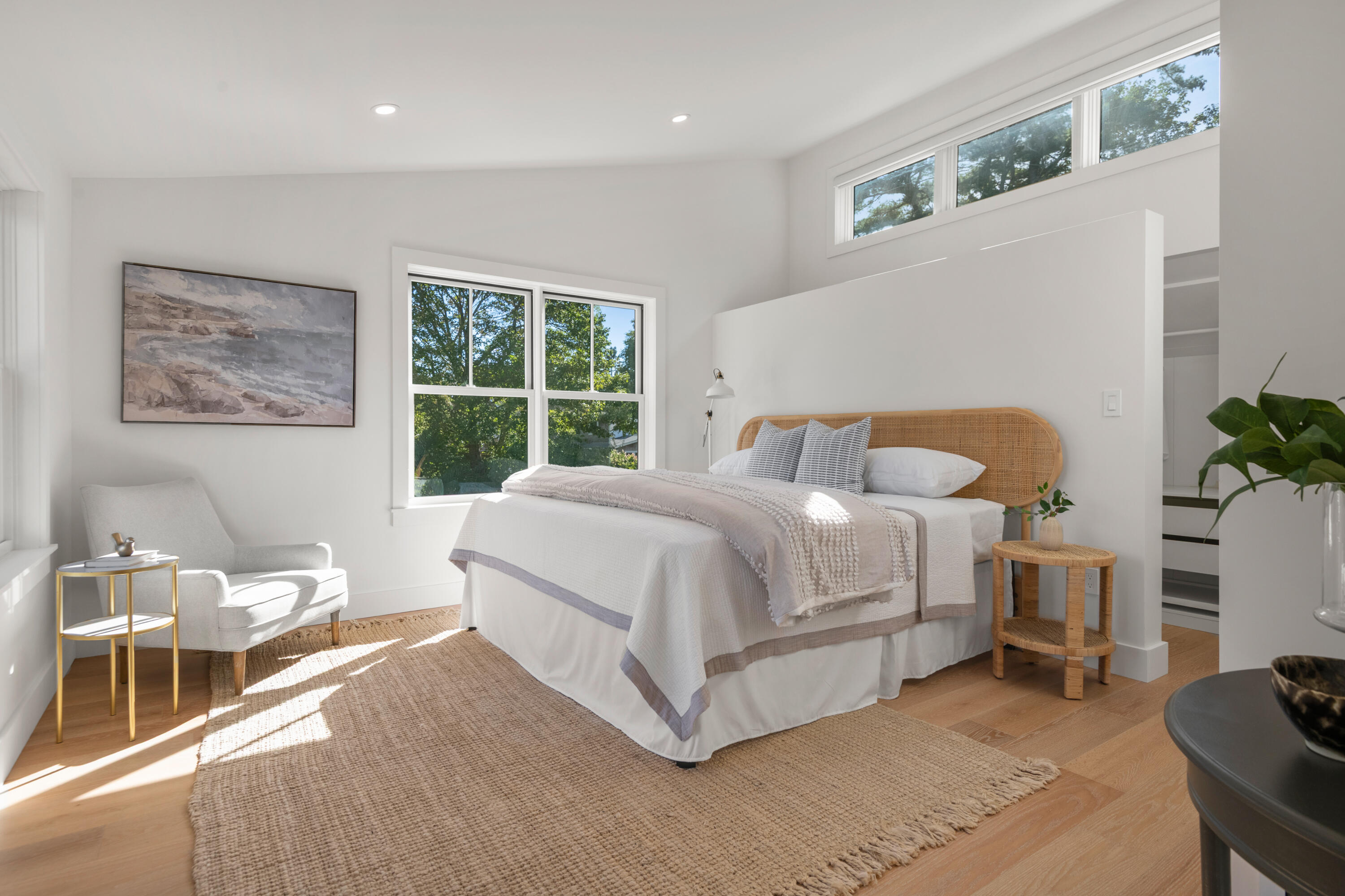 Bright bedroom with a woven-headboard bed, large rug, and tall windows overlooking trees.