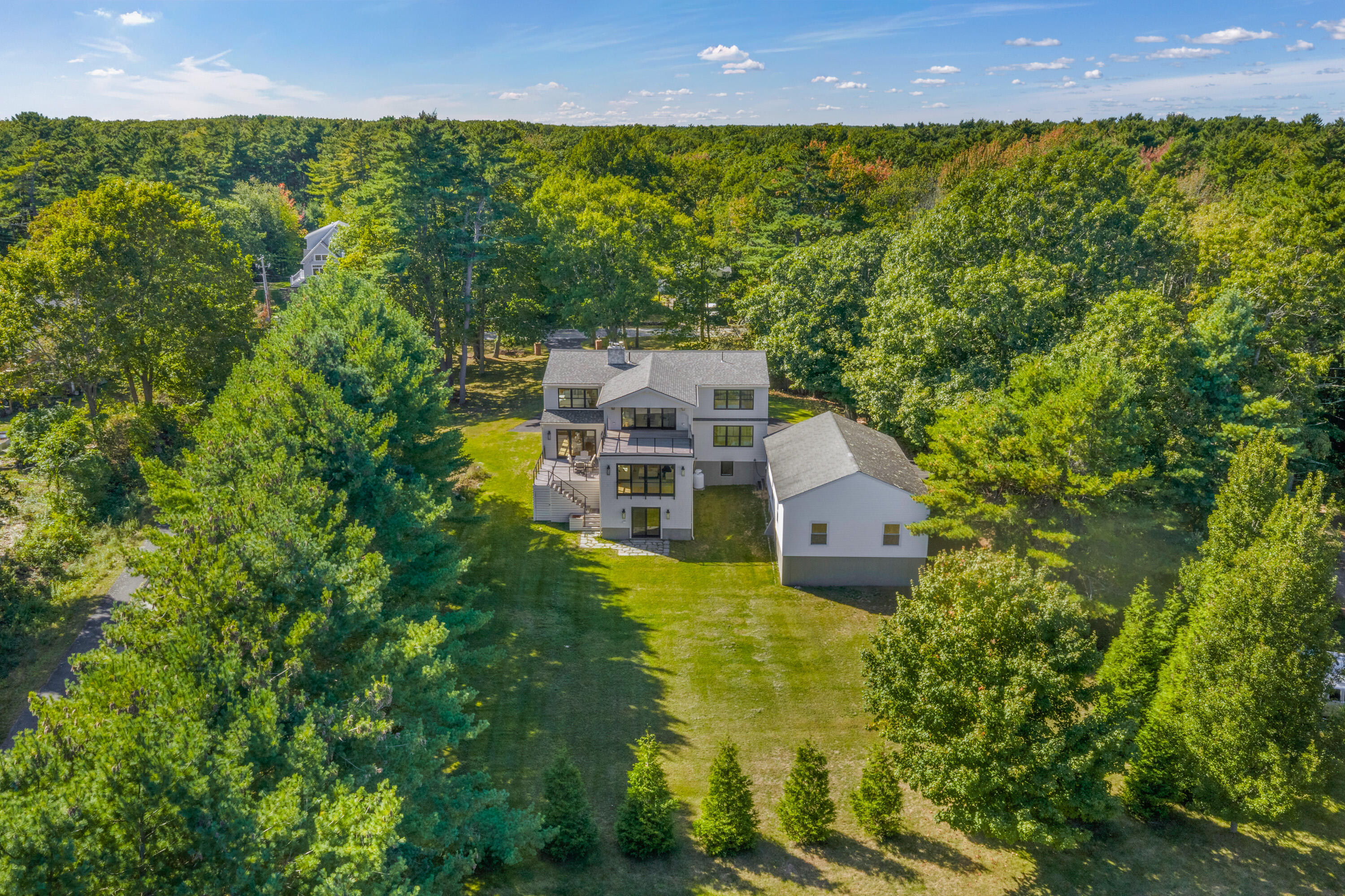 Aerial view of a gray multi-level house with rear decks, a garage, and a wooded lawn.