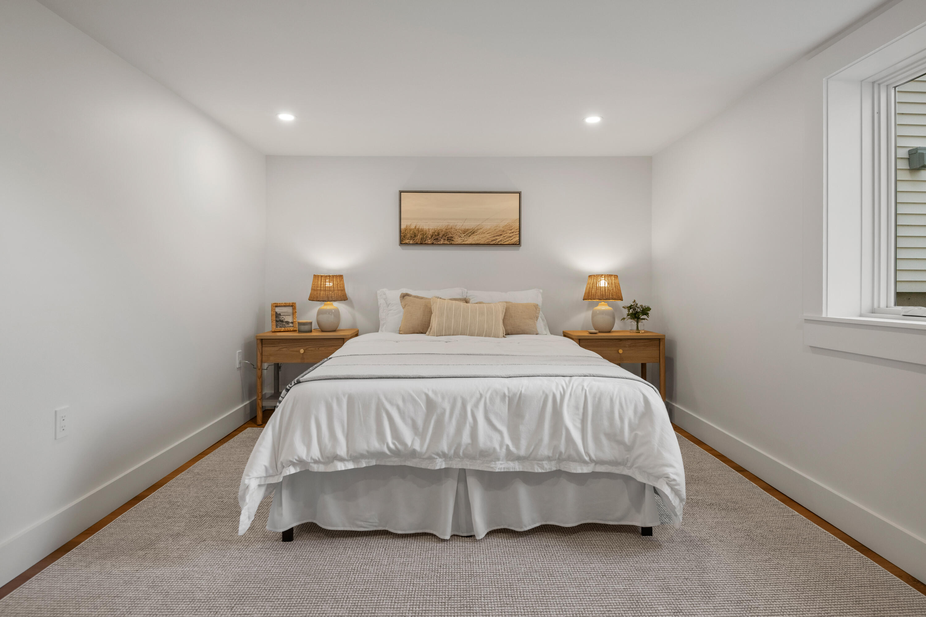 Staged bedroom with a white bed, matching wooden nightstands, and framed coastal grass artwork.