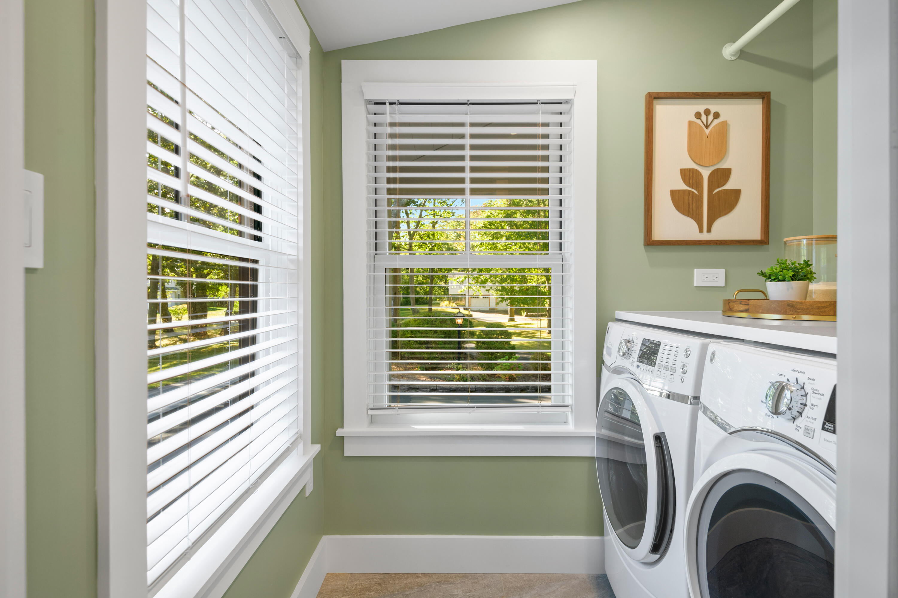 Laundry room with white front-loading washer and dryer beside large windows overlooking a green yard.