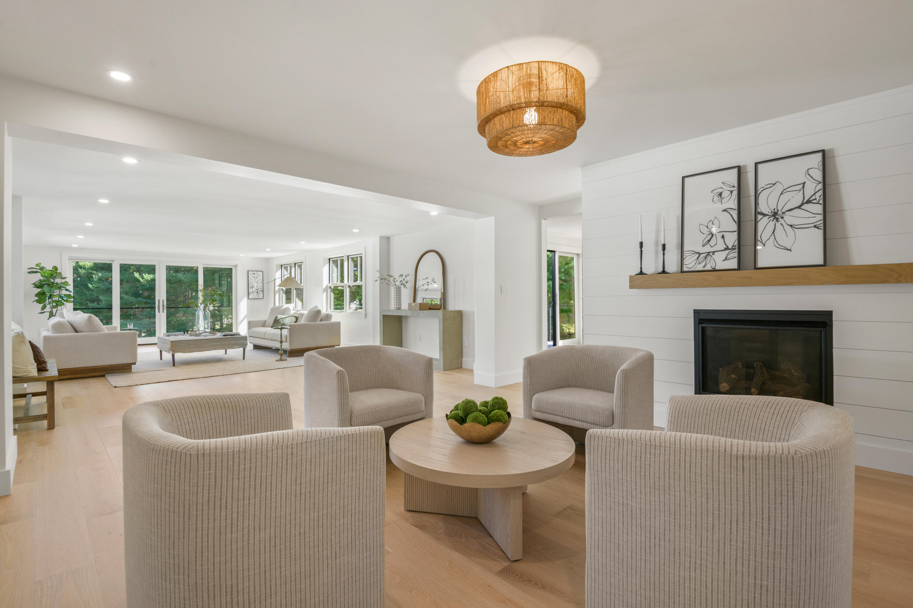 Open-plan living room with a shiplap fireplace, neutral seating, pale wood floors, and large windows.