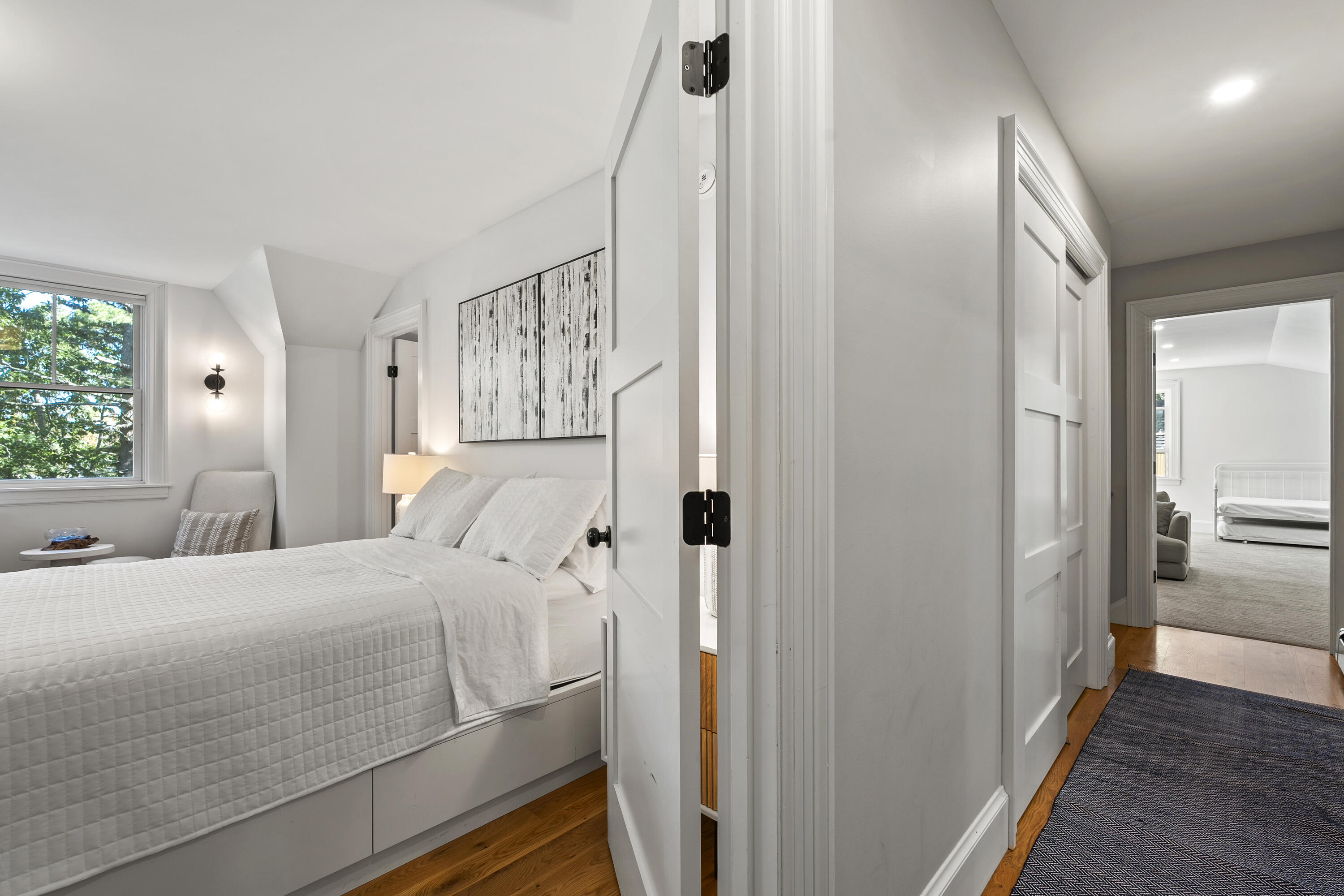 White bedroom with a quilted bed and leafy view through the window, seen from the hallway.