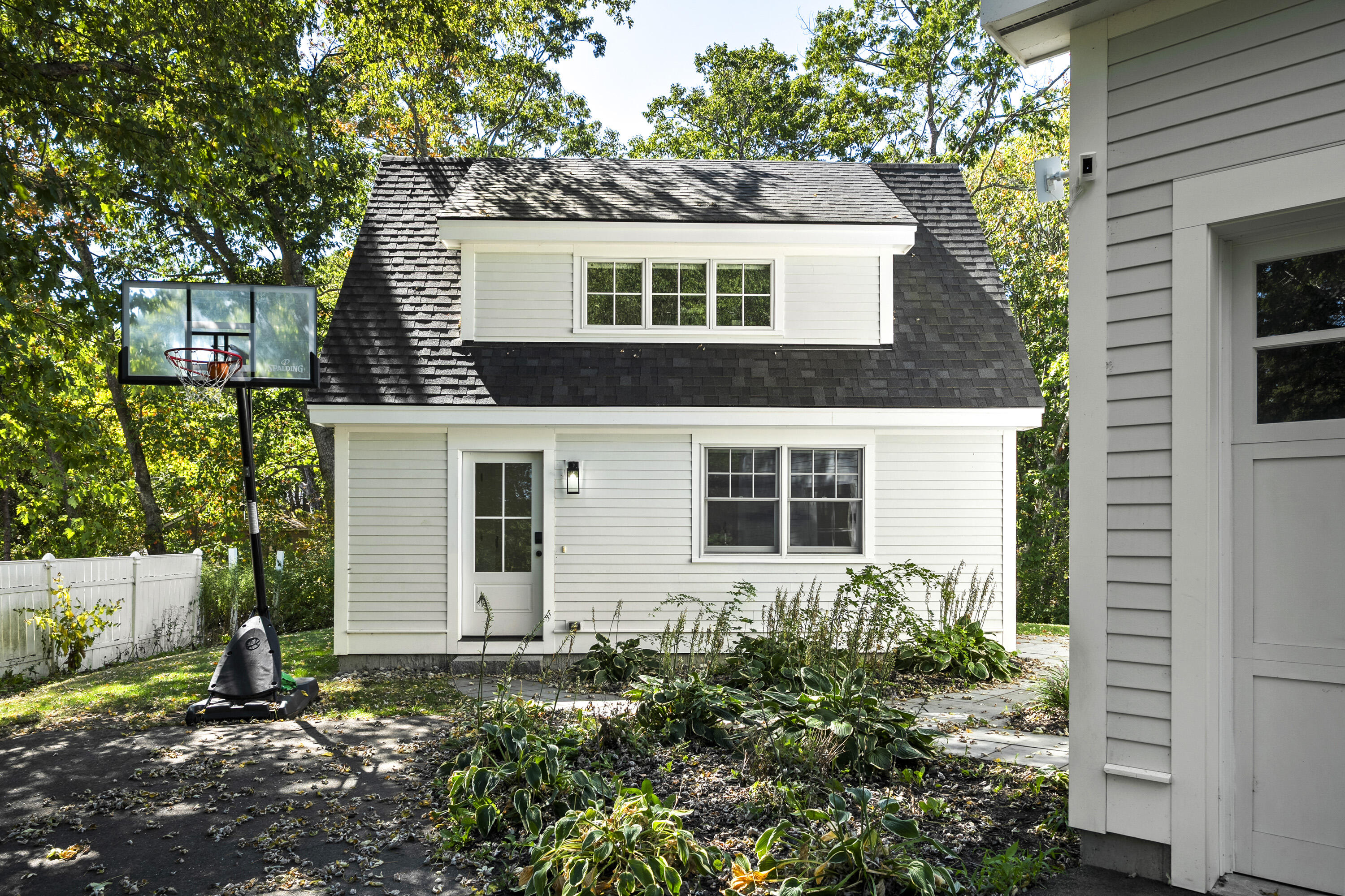 White rear building with a dark shingle roof, dormer, garden beds, and a basketball hoop beside it.