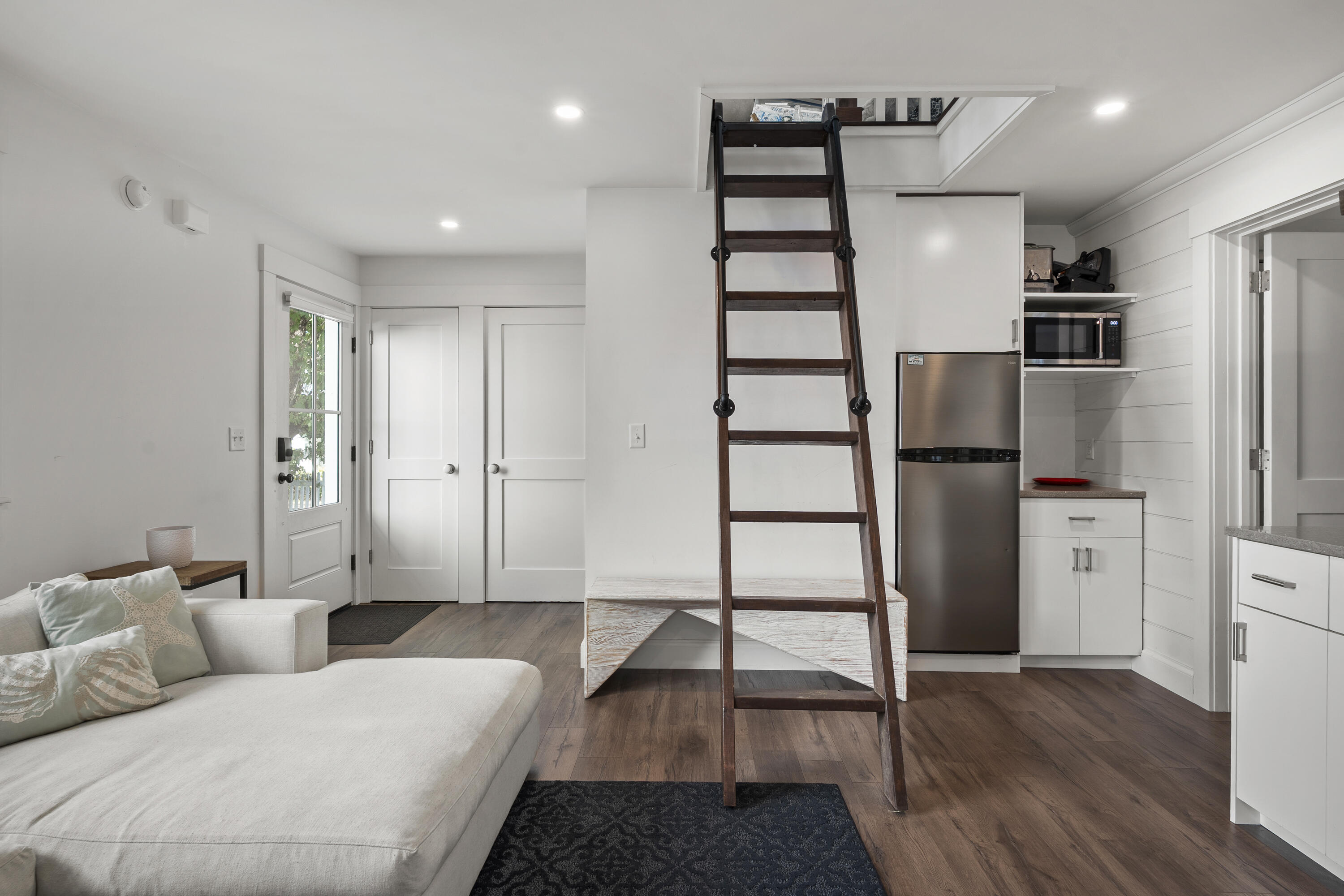 Living room with a white sectional, compact kitchen, and ladder to an overhead loft.
