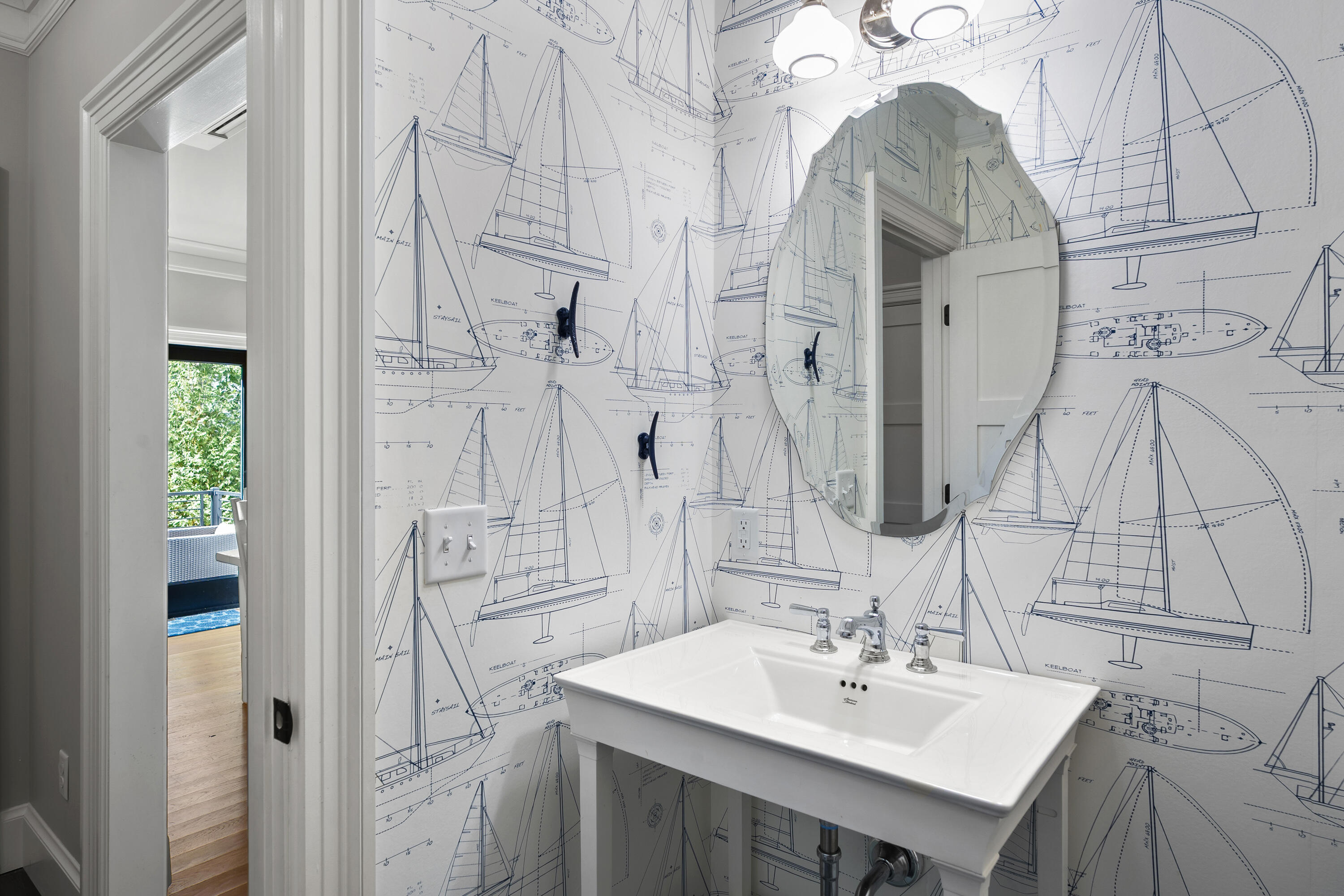 Bathroom with a pedestal sink, oval mirror, and blue sailboat-patterned wallpaper.