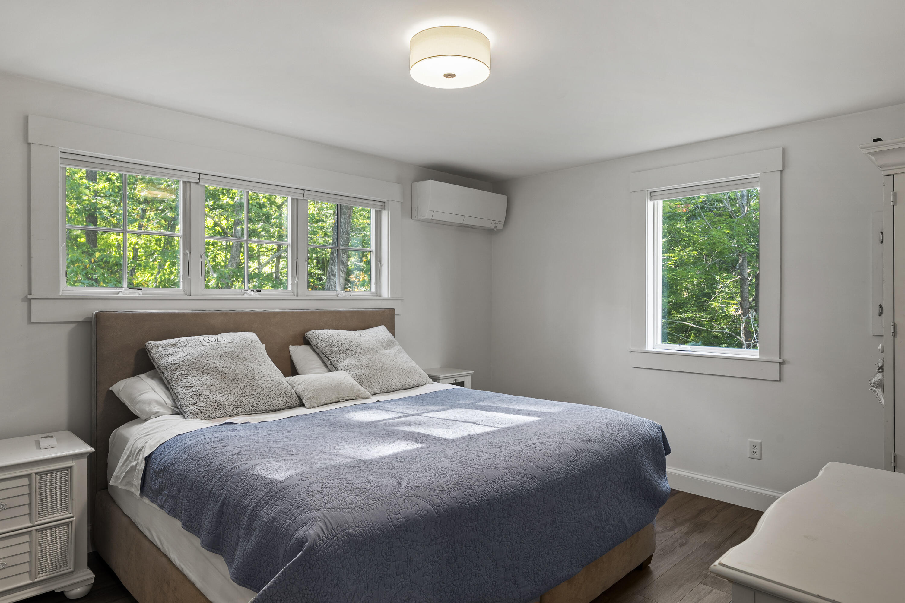 Bedroom with a blue-quilted king bed, large windows overlooking trees, and a wall-mounted mini-split.