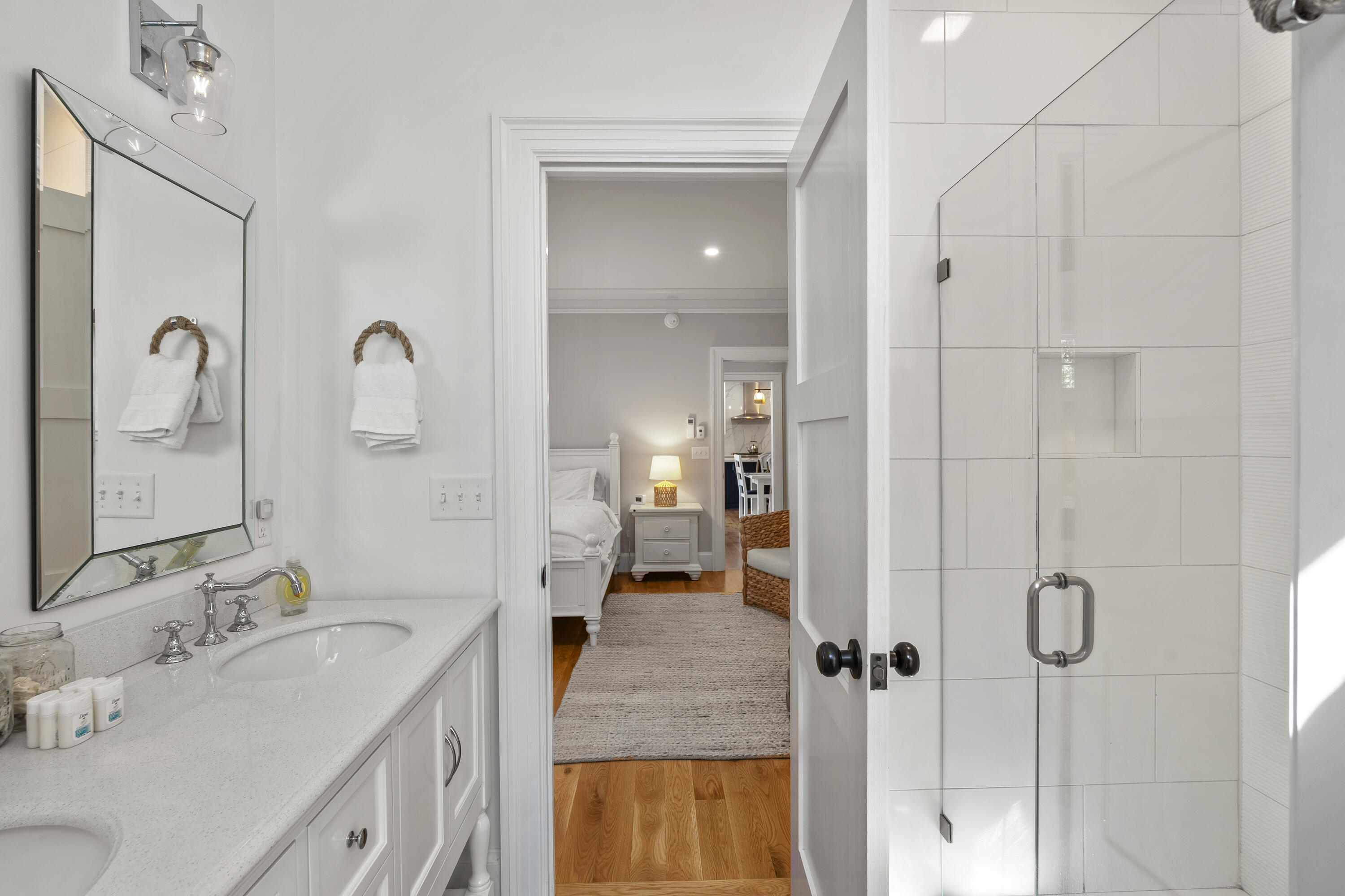 Bathroom with a white vanity and glass shower, looking through an open doorway into a bedroom.
