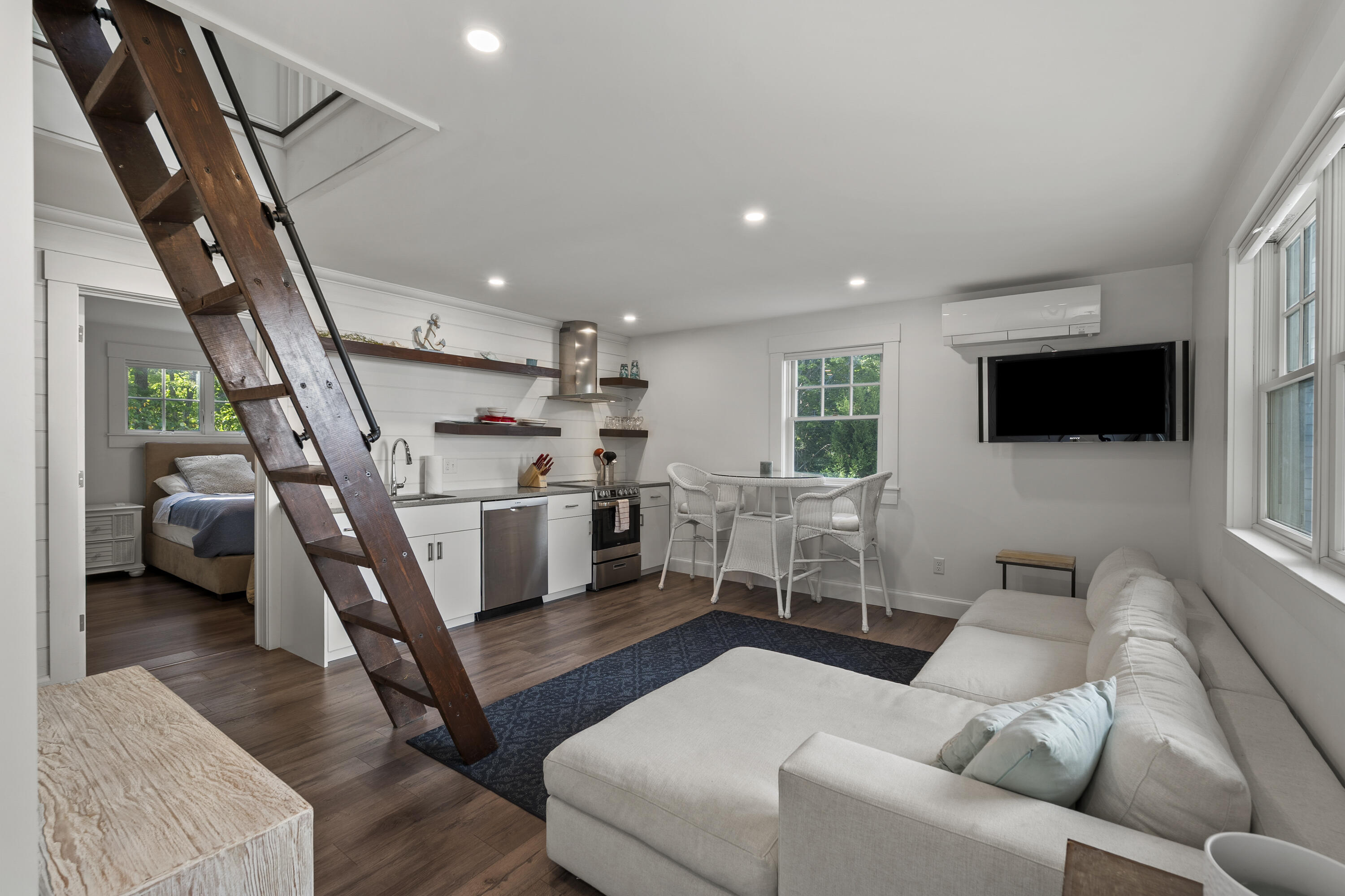 Open-plan studio with a wooden loft ladder, compact kitchen, white sofa, and bedroom nook.