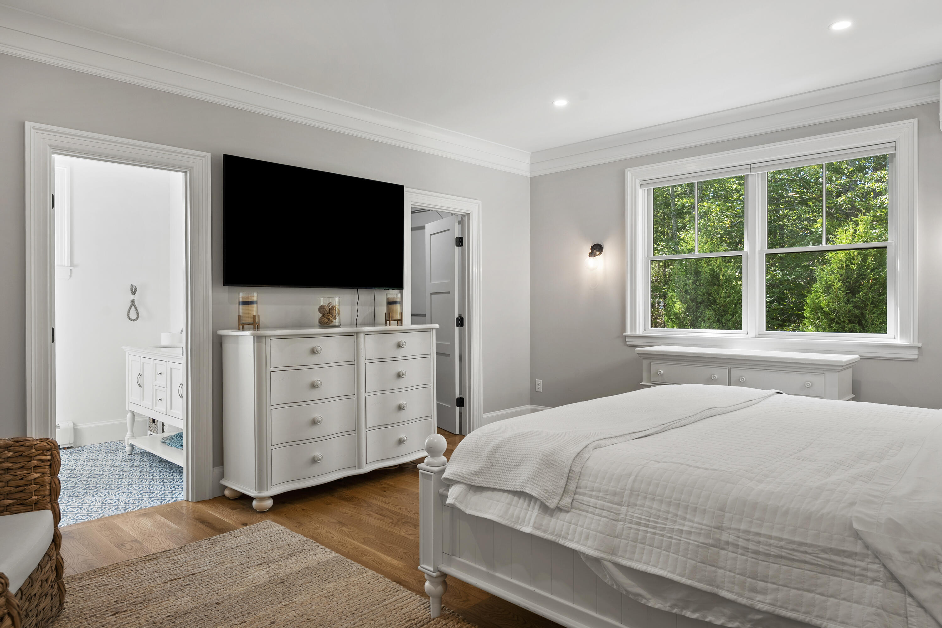 Bright bedroom with a white bed, wall-mounted TV, and large window overlooking green trees.