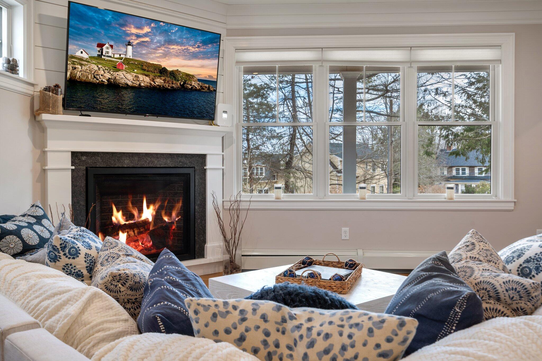 Furnished living room with a lit fireplace, patterned sofas, and large windows overlooking neighboring homes.
