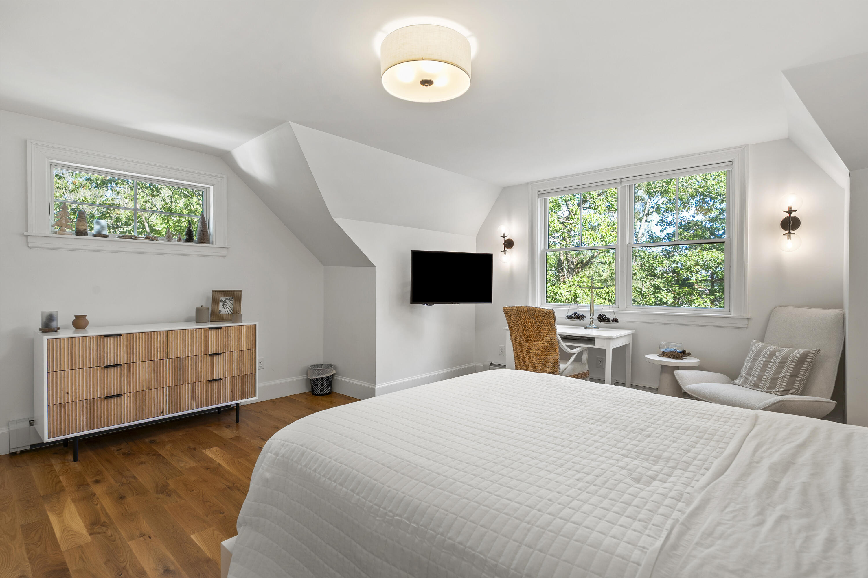 Bedroom with a white quilted bed, fluted wood dresser, and leafy views through wide windows.