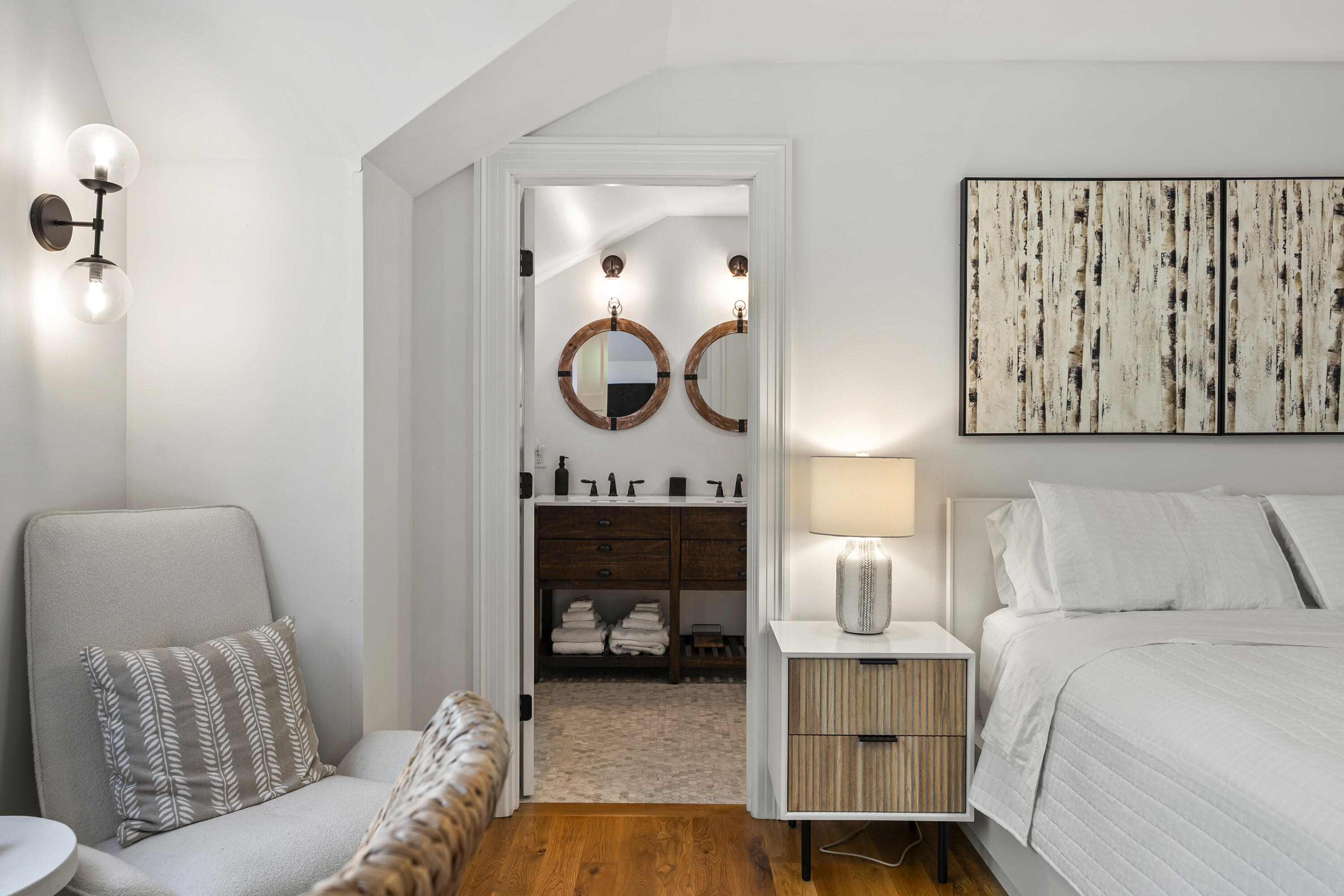 Furnished bedroom with a white bed and chair, opening onto an ensuite bathroom with round mirrors.