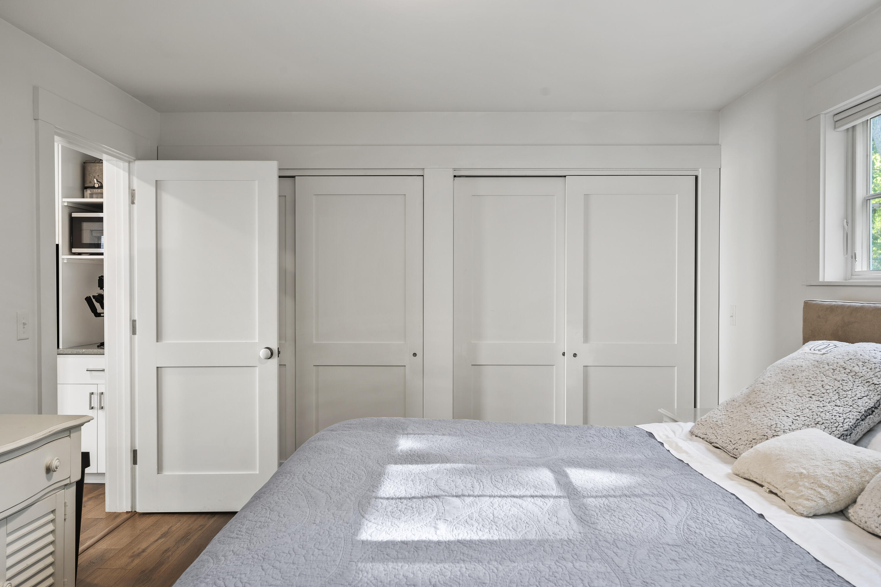 Furnished bedroom with a gray quilted bed, white sliding doors, and a sunlit window.