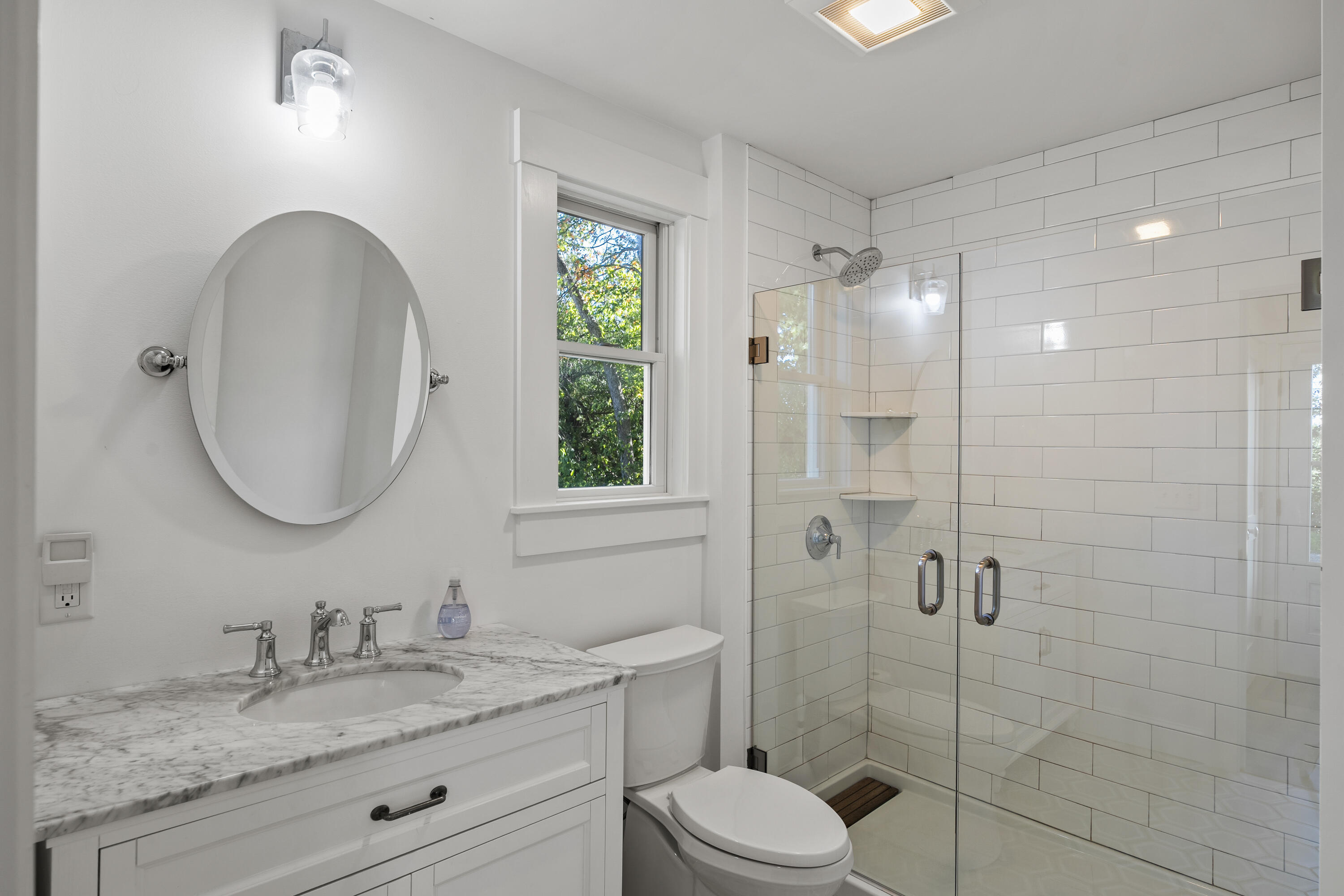 Bathroom with marble vanity and glass-enclosed subway-tile shower beside a leafy-view window.