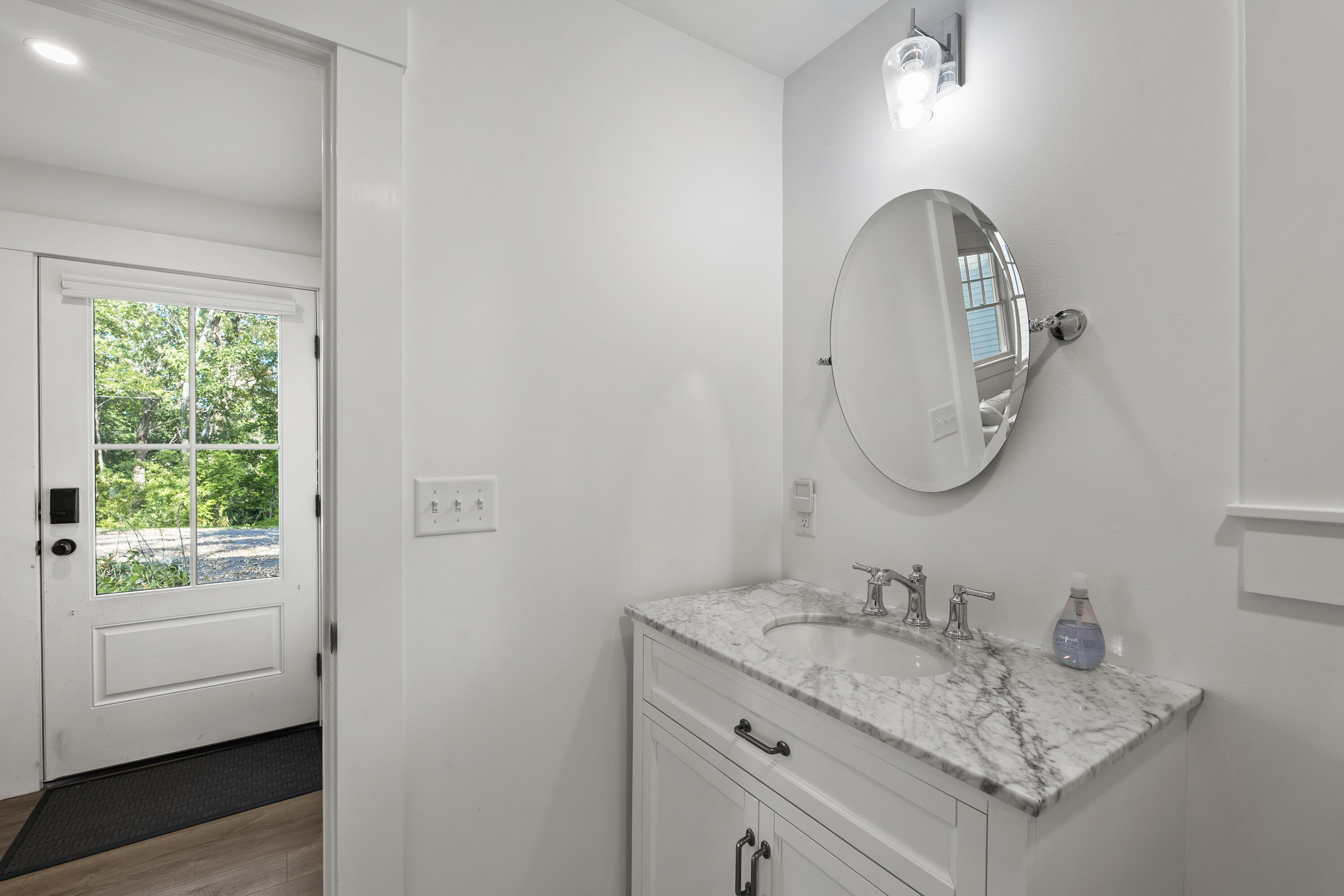 White bathroom vanity with marble-pattern counter and round mirror beside a glass-paneled door.