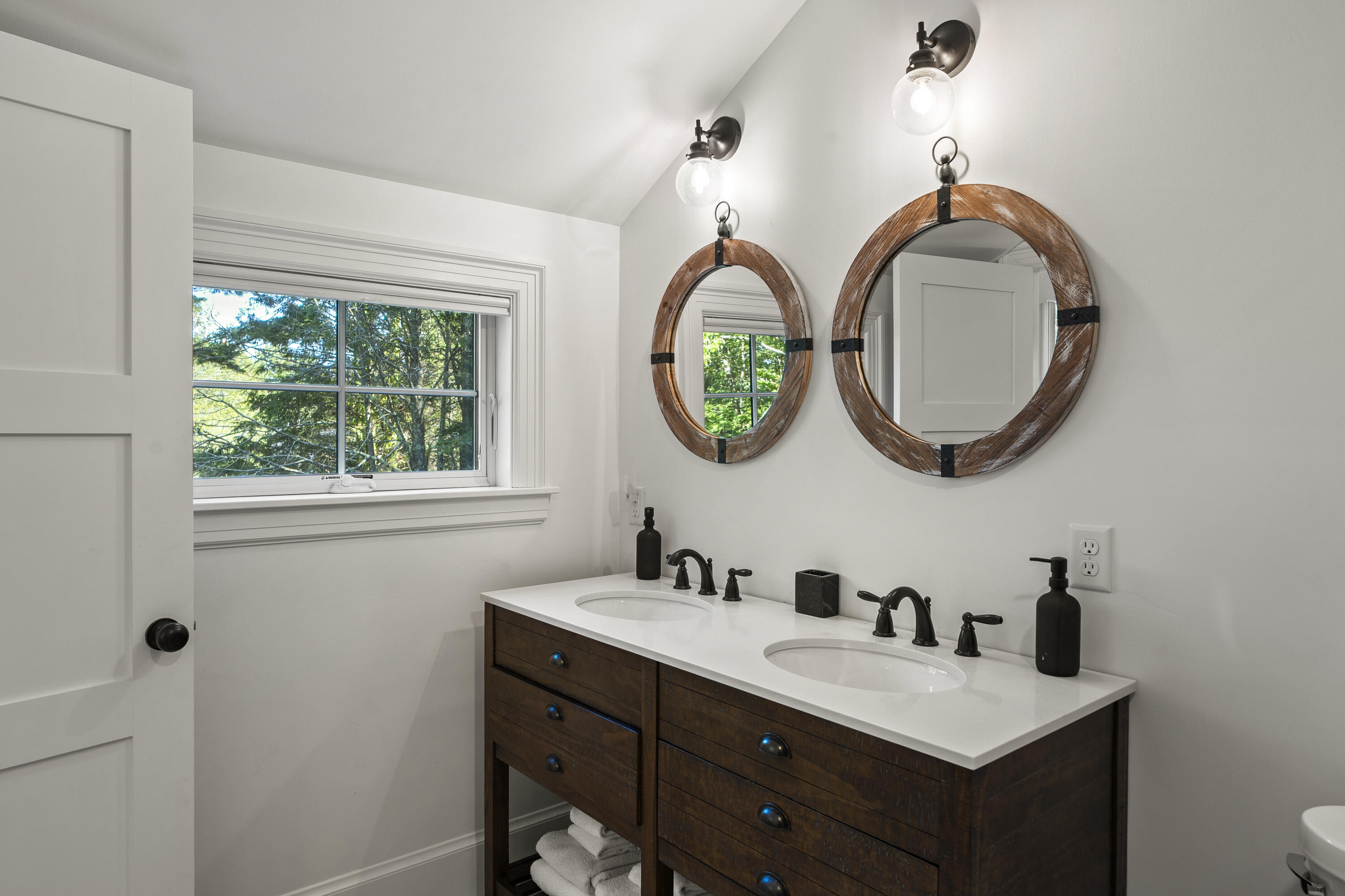 Bathroom with a dark wood double vanity, paired round mirrors, and a window overlooking trees.