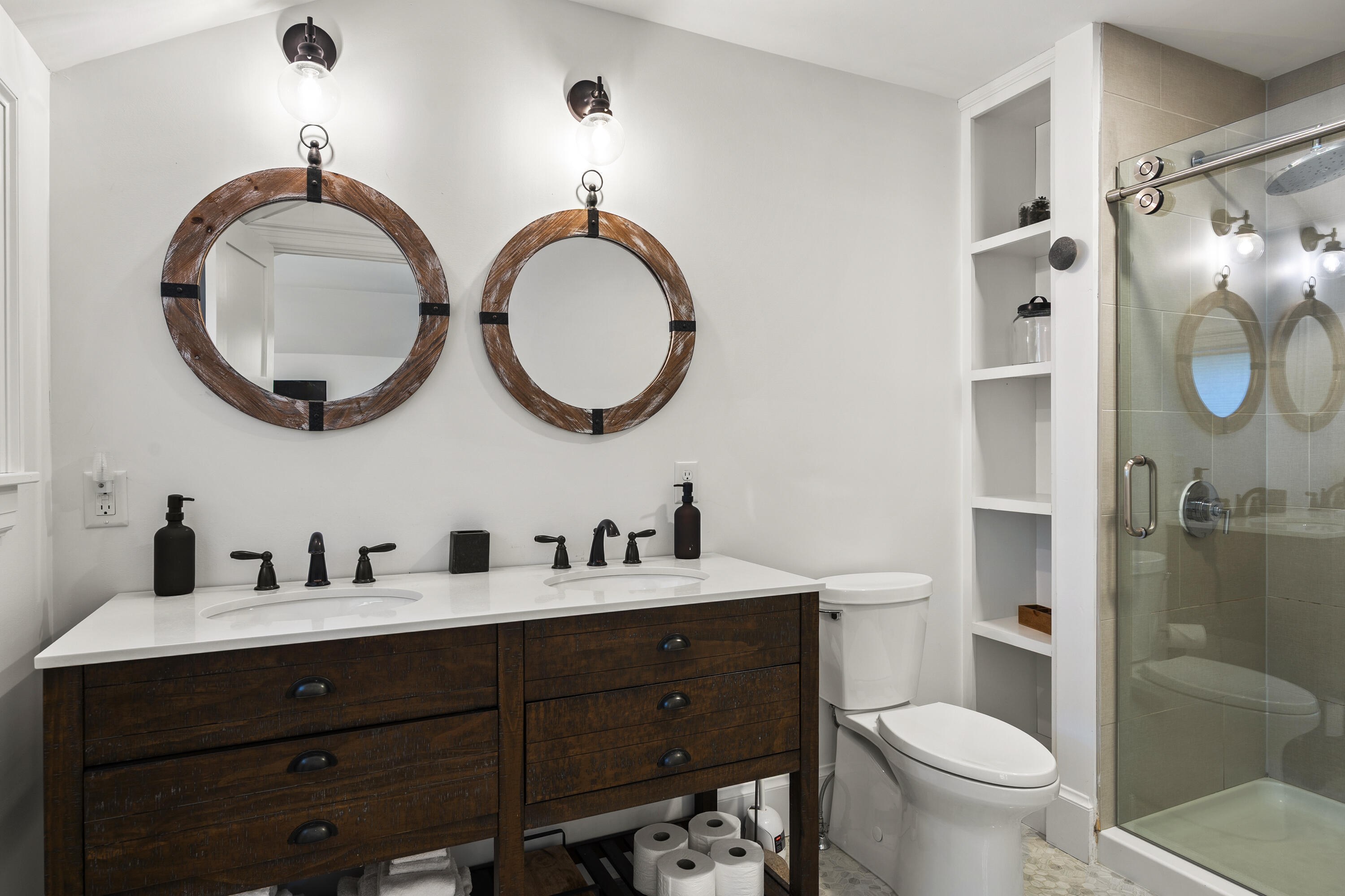 Bathroom with a dark wood double vanity, round mirrors, built-in shelves, and glass-enclosed shower.