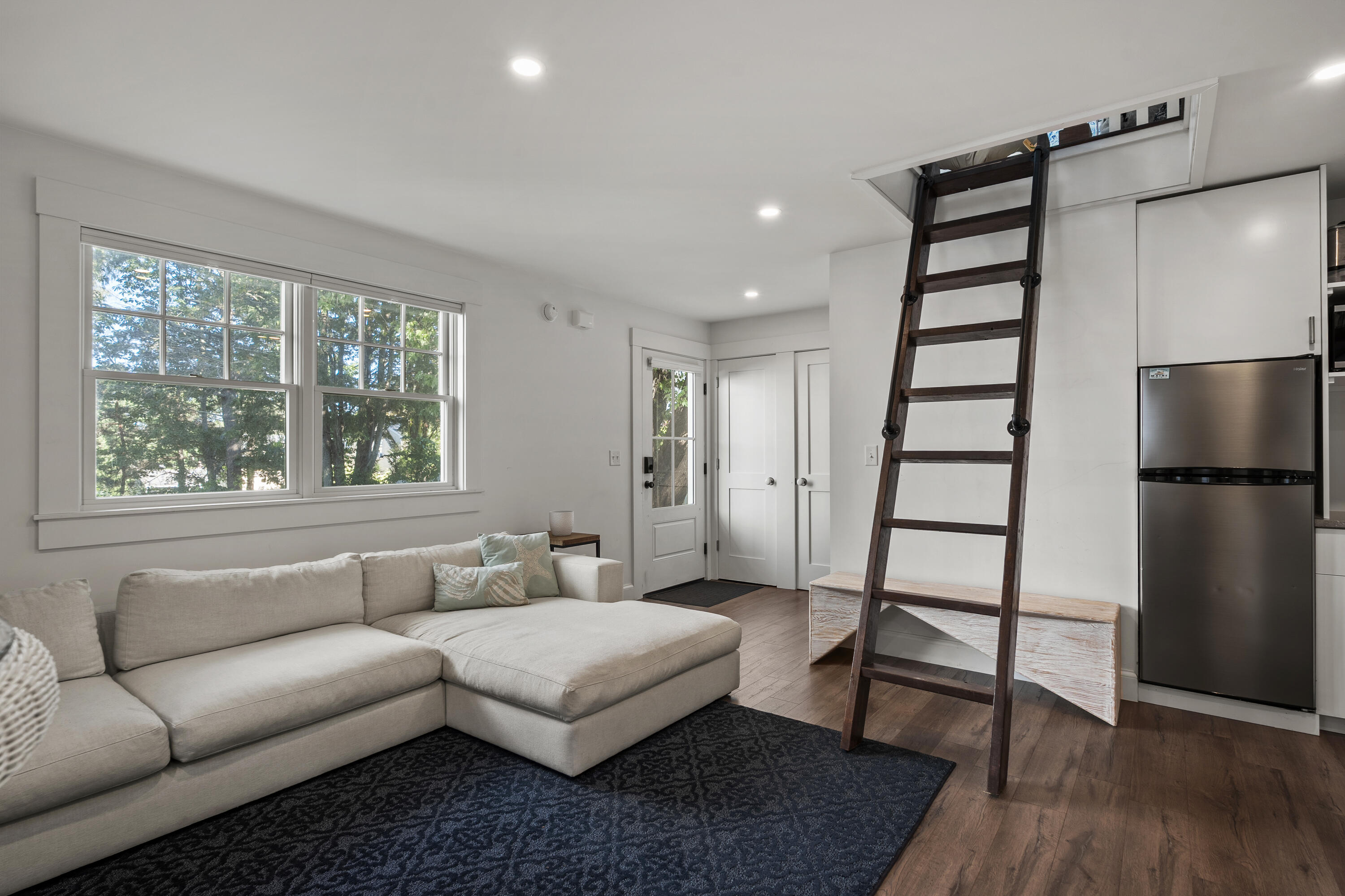 Living area with a light sectional, large tree-facing windows, and a ladder to a ceiling hatch.