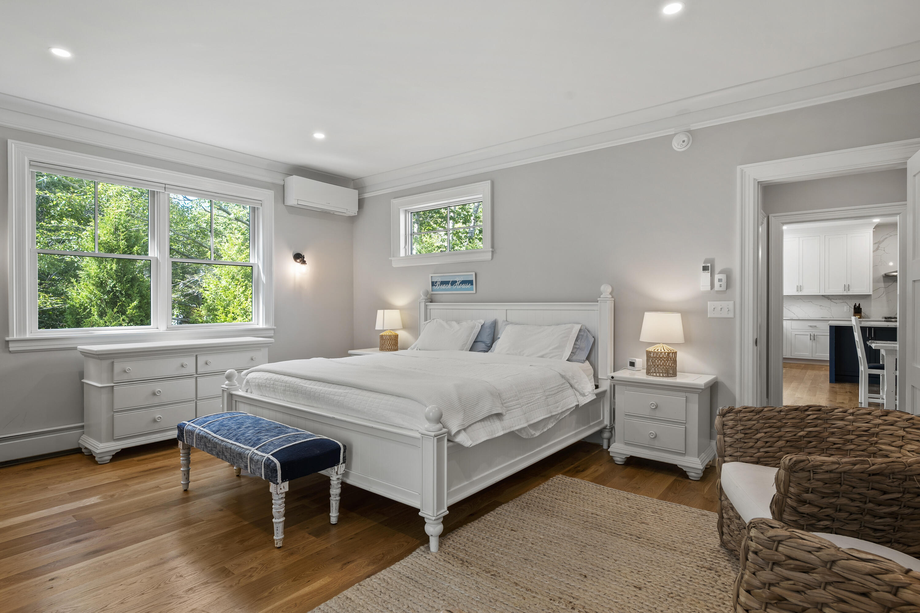 Bright bedroom with a white bed, blue bench, and leafy views through large windows.