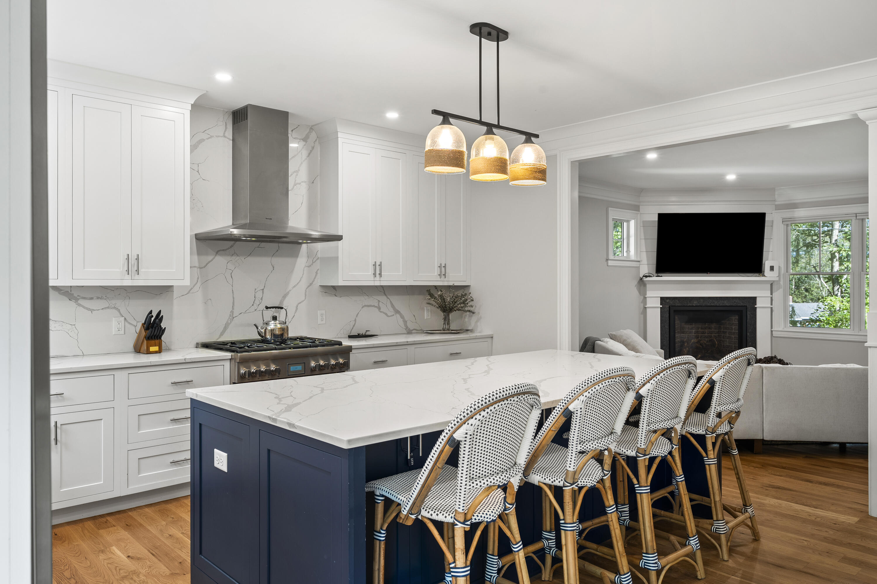 Kitchen with a navy island, veined stone surfaces, woven bar stools, and an adjoining living room with a fireplace.
