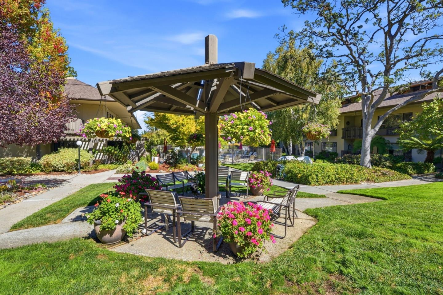 Residential courtyard with a wooden gazebo, patio seating, colorful flowers, and a pool in the background.