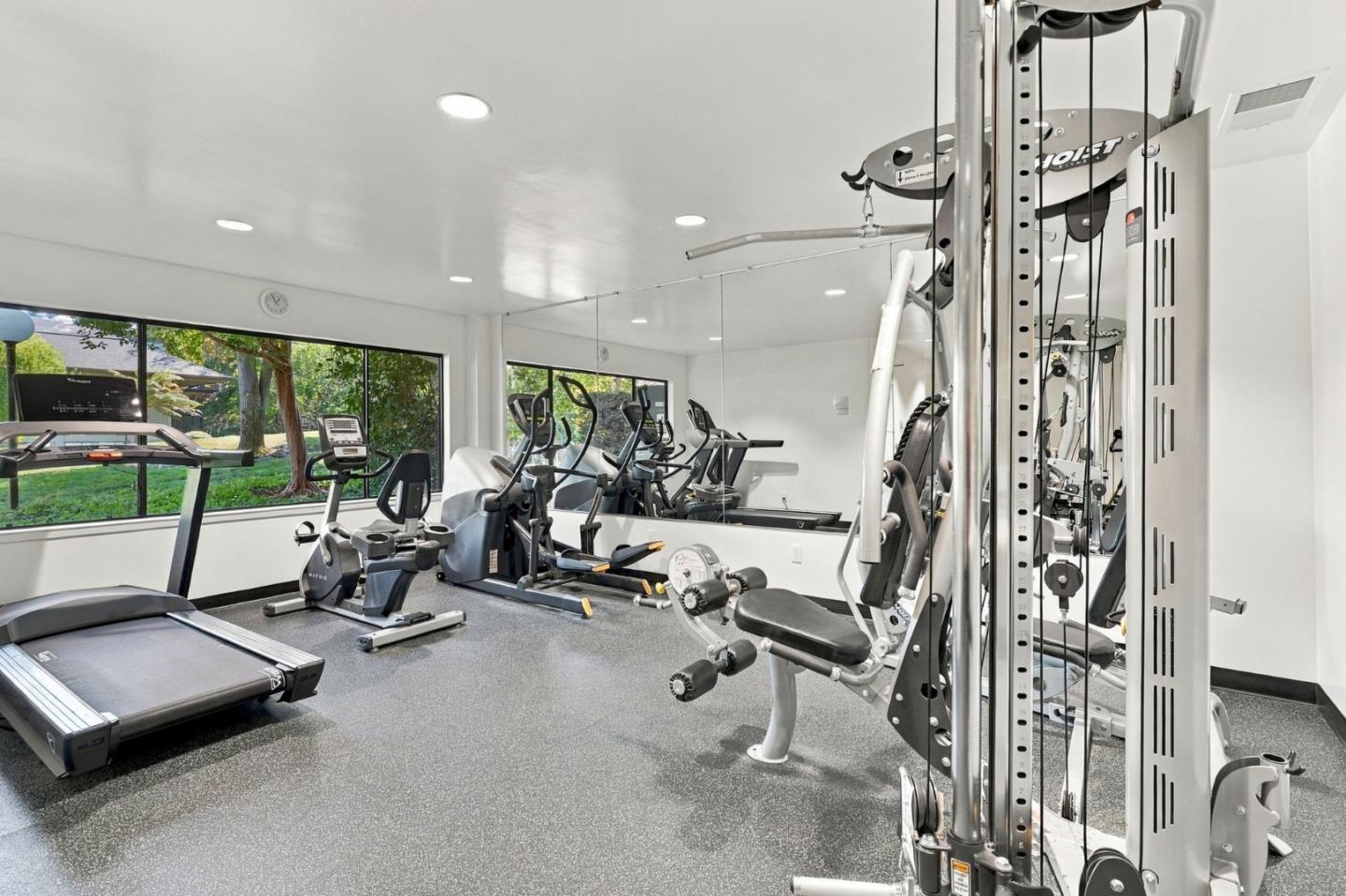 Fitness room with treadmills, stationary bikes, and weight machines beside large windows.