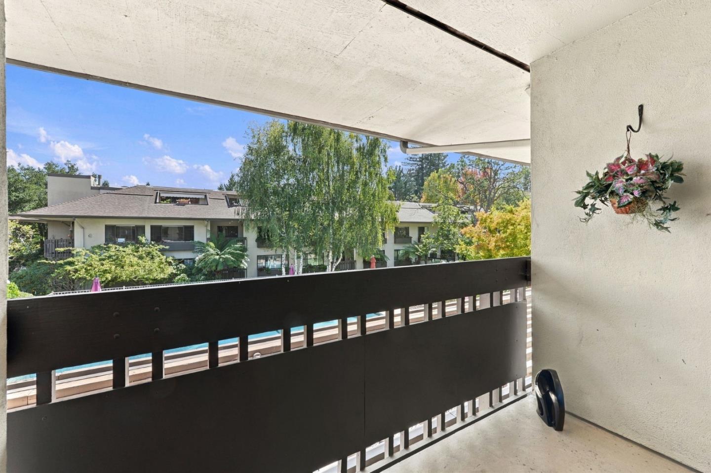 Covered apartment balcony overlooking a swimming pool, trees, and a neighboring residential building.