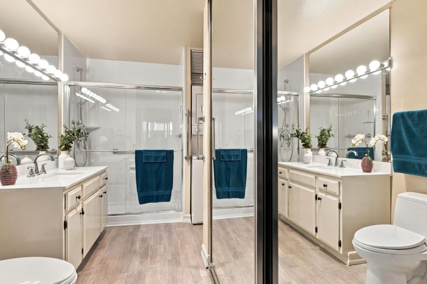 Bathroom with a glass shower, mirrored white vanity, bright bulb lighting, and teal towels.