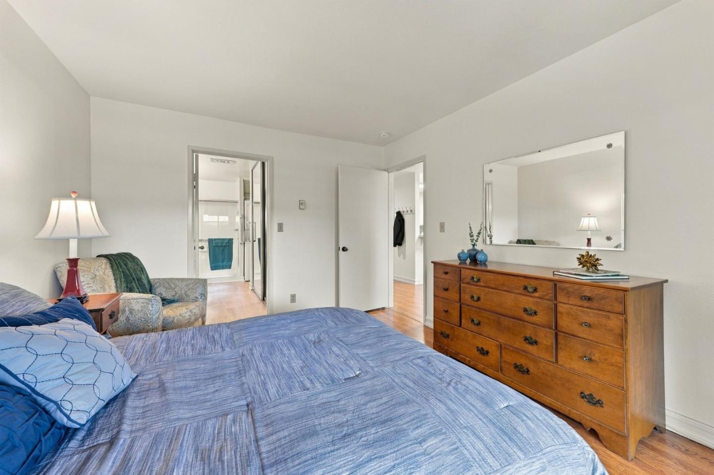 Furnished bedroom with a blue quilted bed, patterned armchair, and wooden dresser beneath a large mirror.