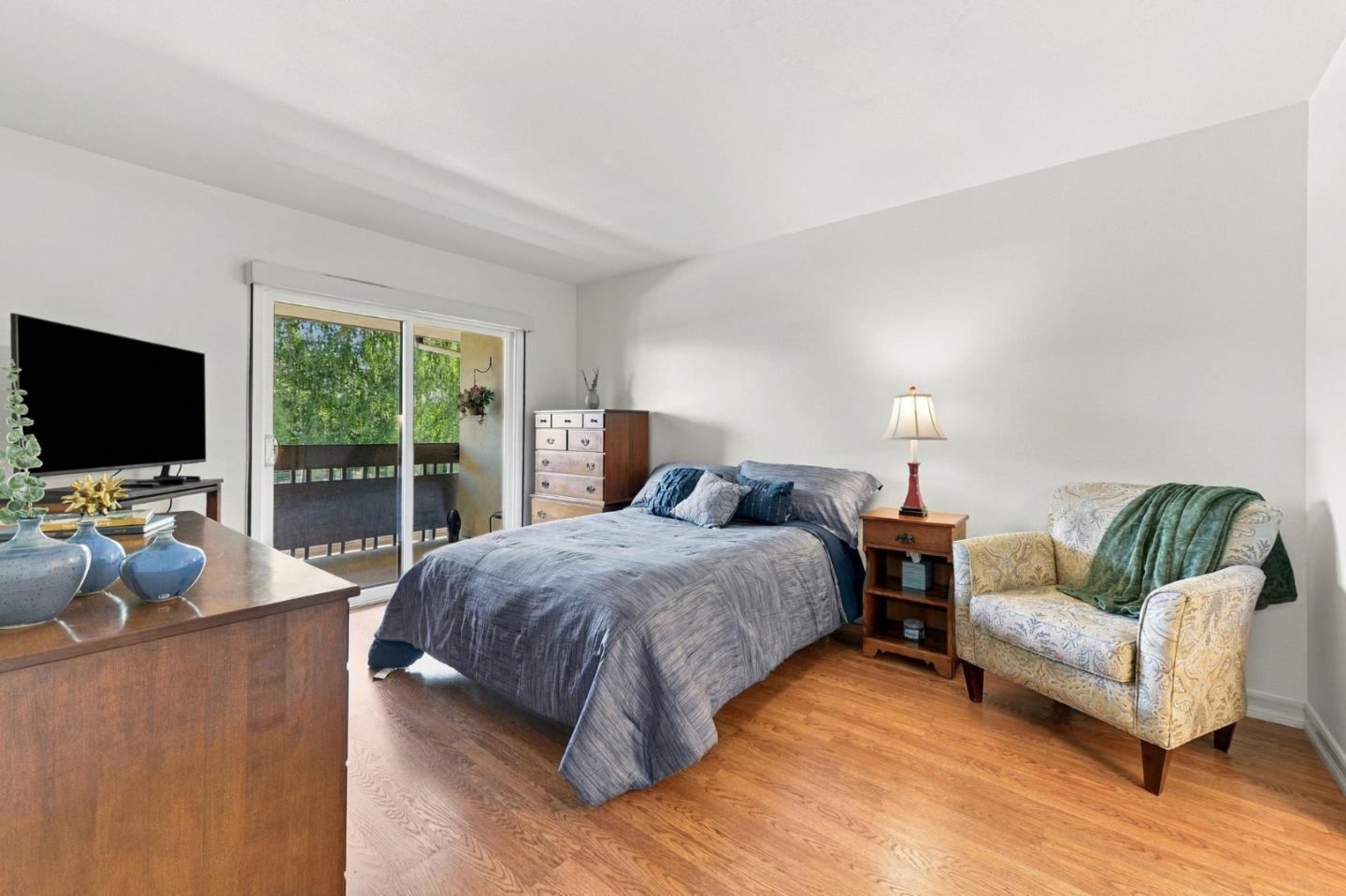 Bedroom with gray bedding, hardwood floors, and sliding doors to a leafy balcony.