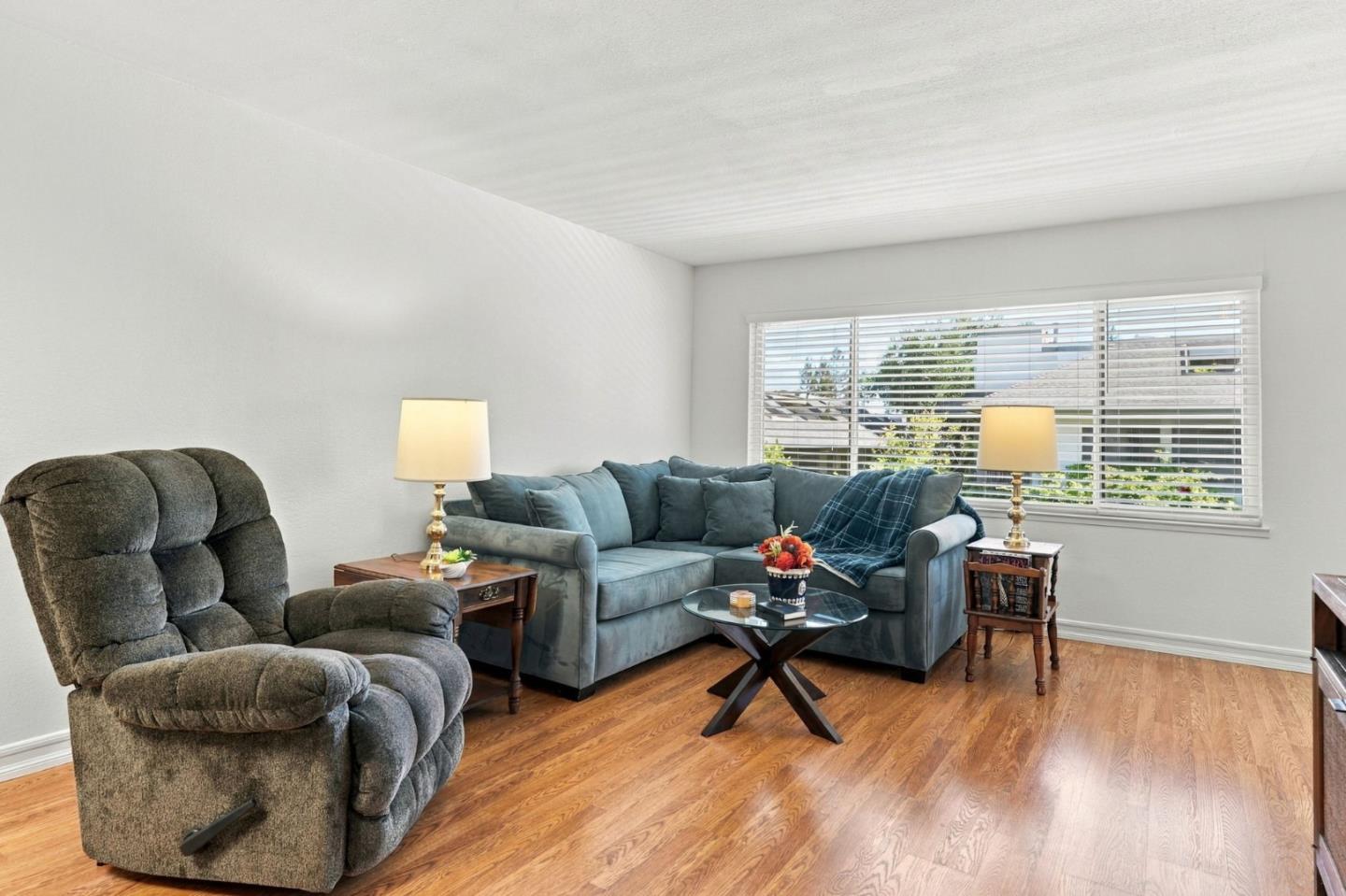 Living room with a blue sectional, gray recliner, glass coffee table, and wide window with blinds.