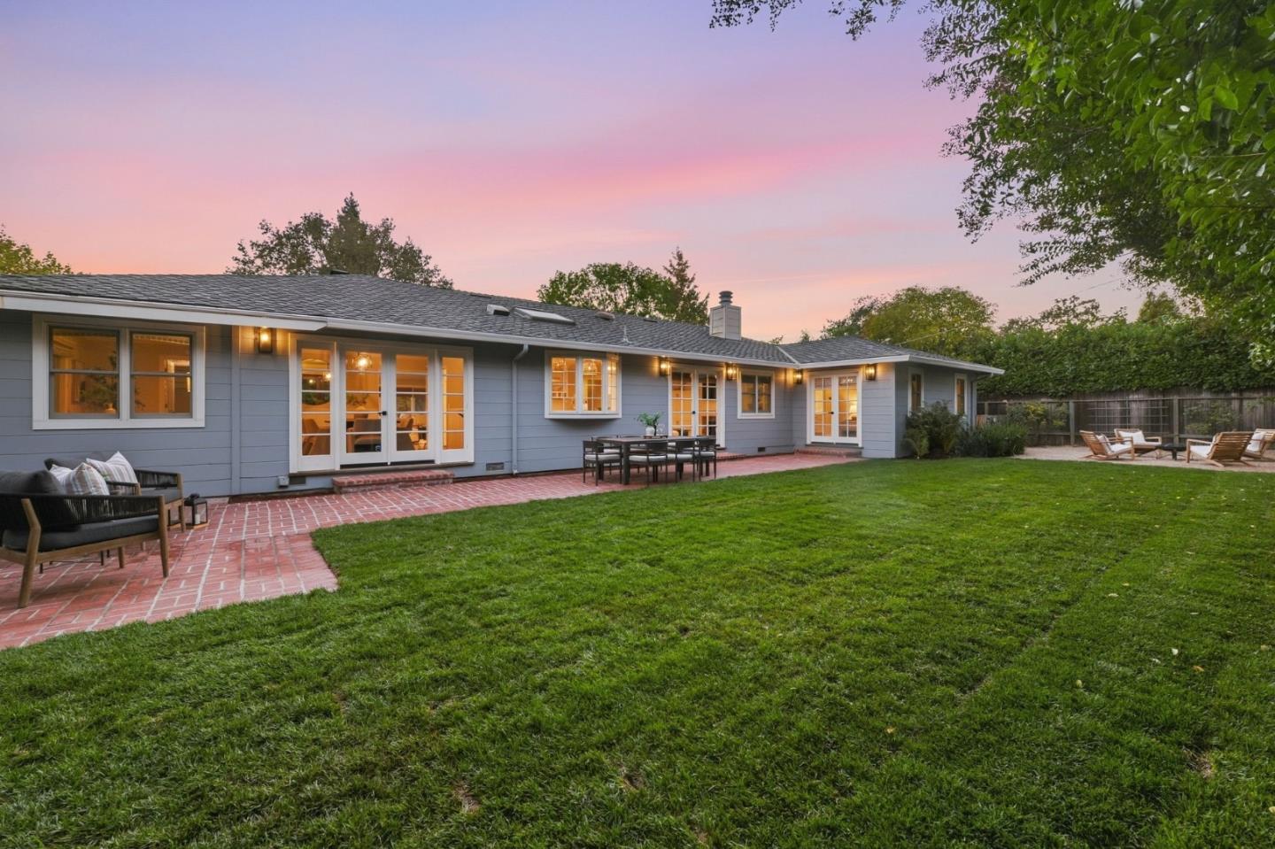 Rear of a gray home with a brick patio, broad lawn, and outdoor seating at dusk.