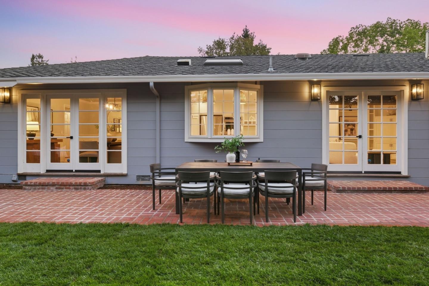 Gray home’s rear exterior with French doors, brick patio, and dining set at dusk.