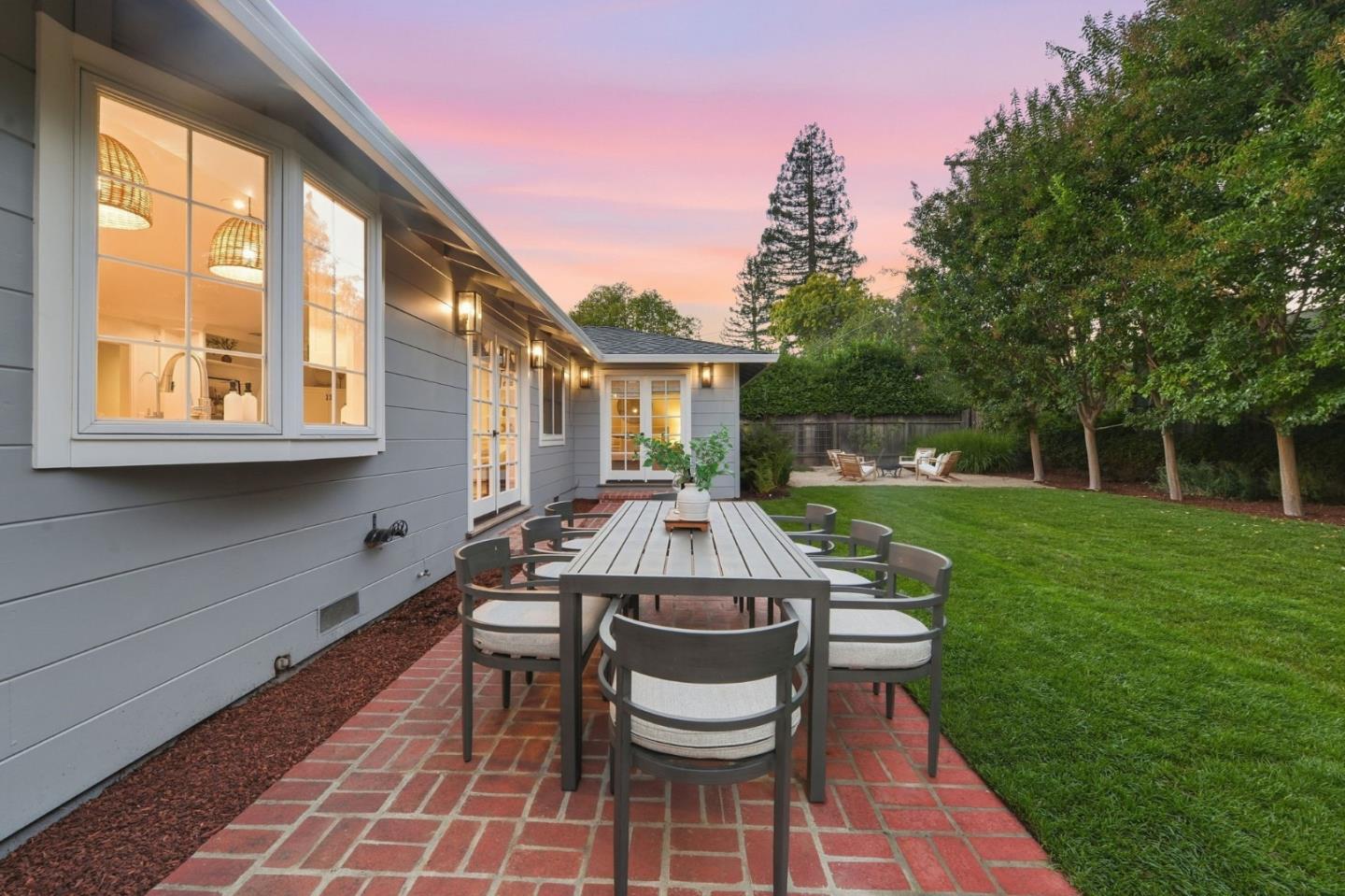 Brick patio dining area beside a gray home, with a lawn and mature trees at dusk.