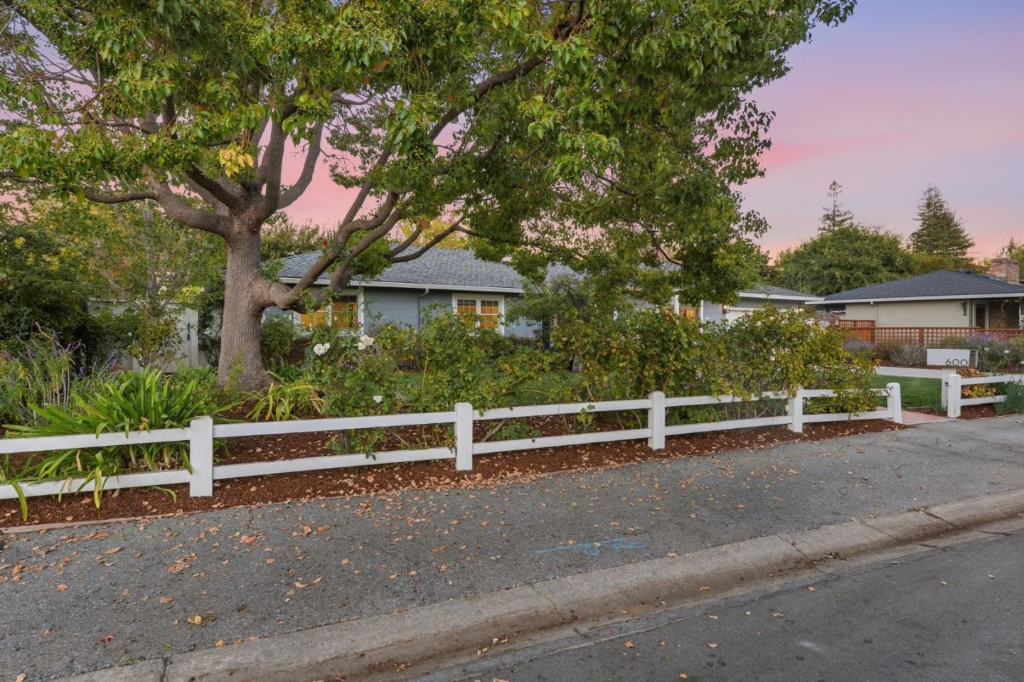 Single-story home behind a white fence and mature tree at dusk.
