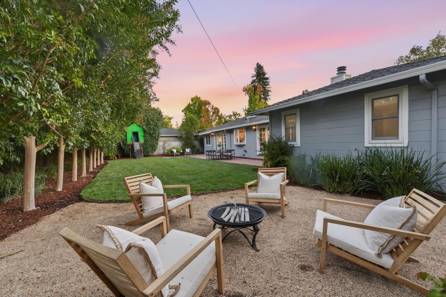 Landscaped backyard with a gravel fire-pit seating area, lawn, and green playhouse at dusk.