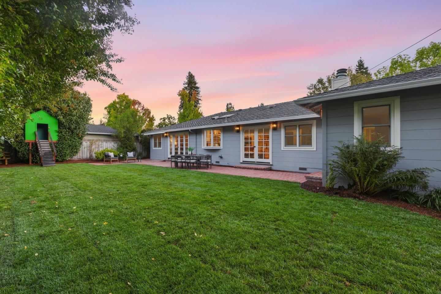 Gray home's backyard with a broad lawn, brick patio, and green playhouse at dusk.