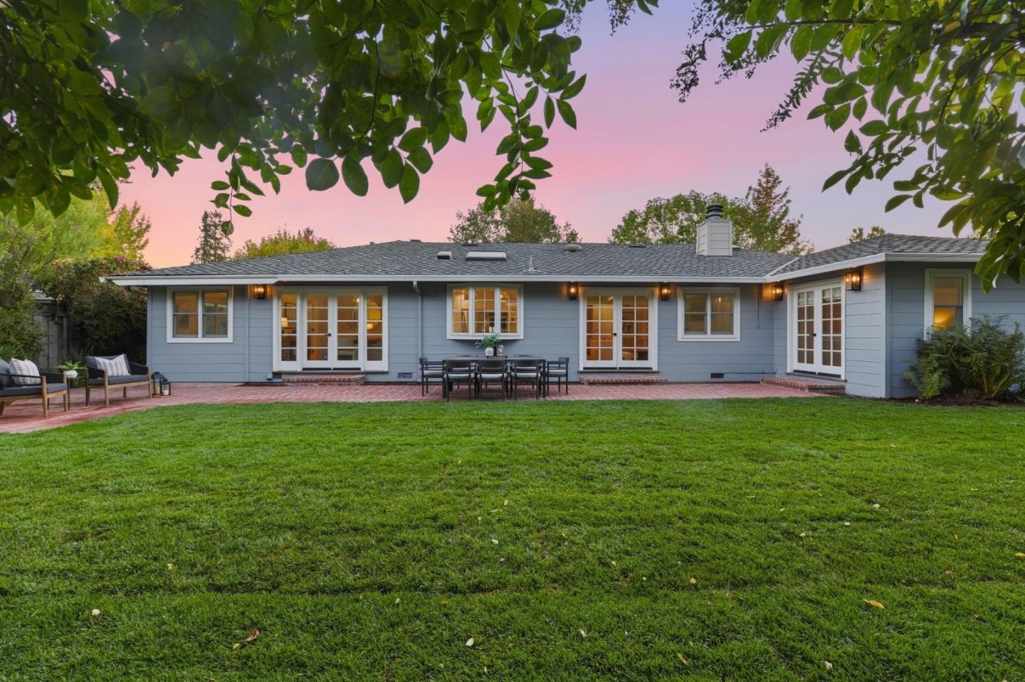 Rear of a blue-gray single-story home with French doors, brick patio, lawn, and outdoor dining area at dusk.