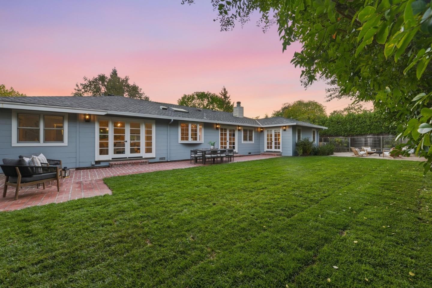 Blue single-story home’s rear facade and brick patio overlook a broad lawn at dusk.