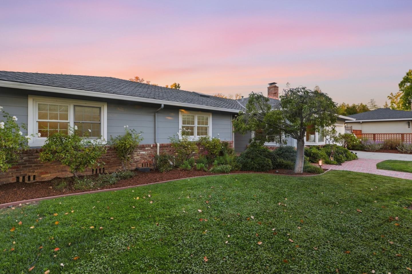 Single-story gray house with a brick lower facade, landscaped lawn, and curved brick walkway at dusk.
