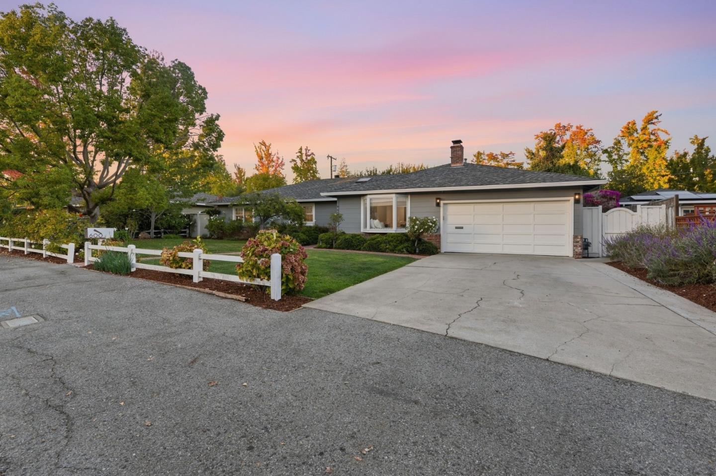 Single-story gray home with a wide garage, landscaped lawn, white fence, and driveway at dusk.