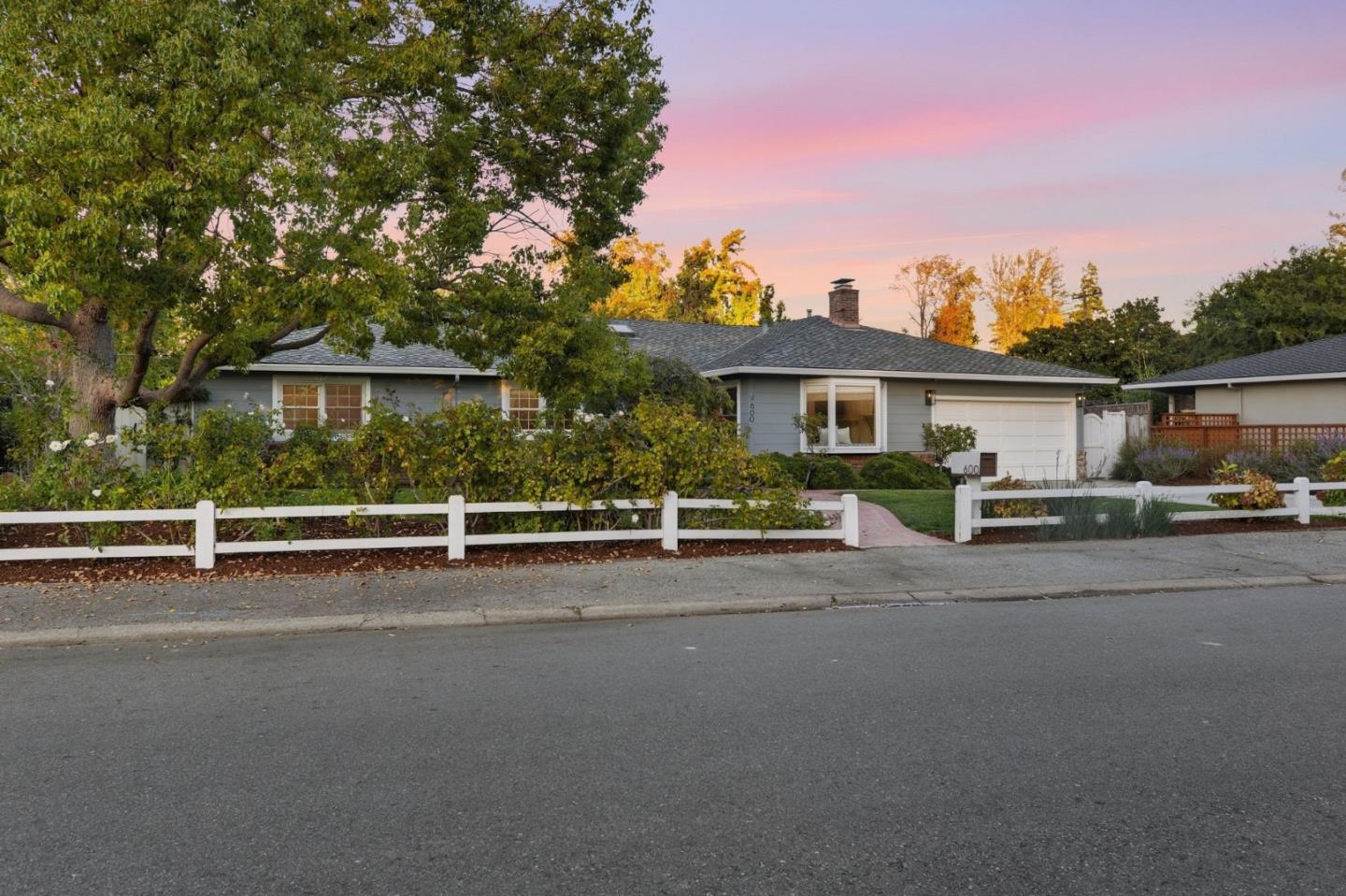 Single-story home behind a white fence, framed by mature trees beneath a pink dusk sky.