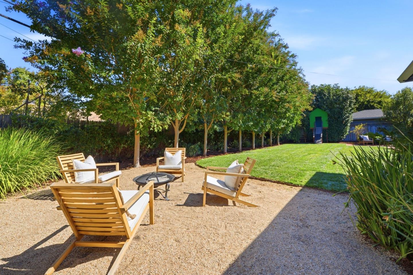 Backyard seating area with wooden chairs, a fire pit, lawn, mature trees, and a green play structure.