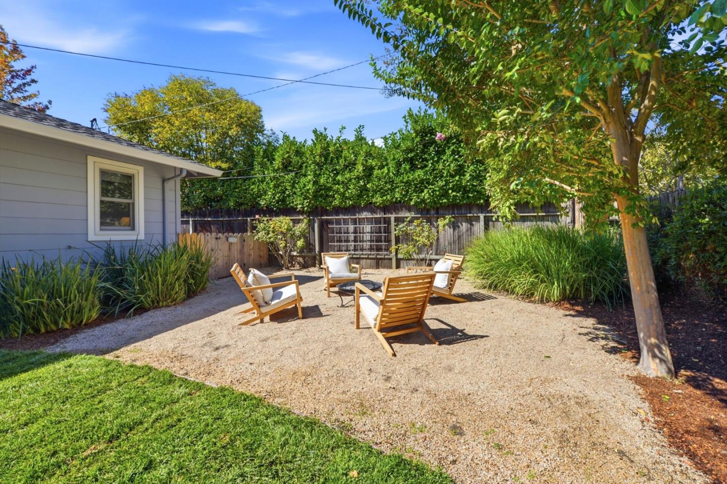 Backyard gravel patio with wooden lounge chairs around a fire pit beneath leafy trees.