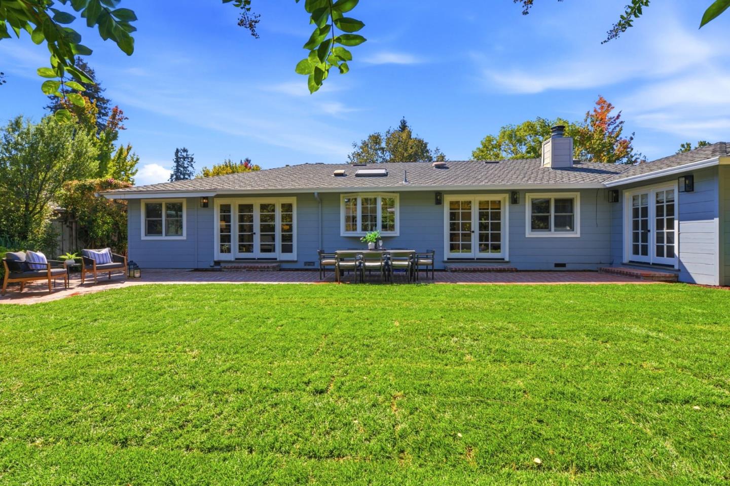 Blue single-story home behind a broad lawn, with a brick patio and outdoor dining area.