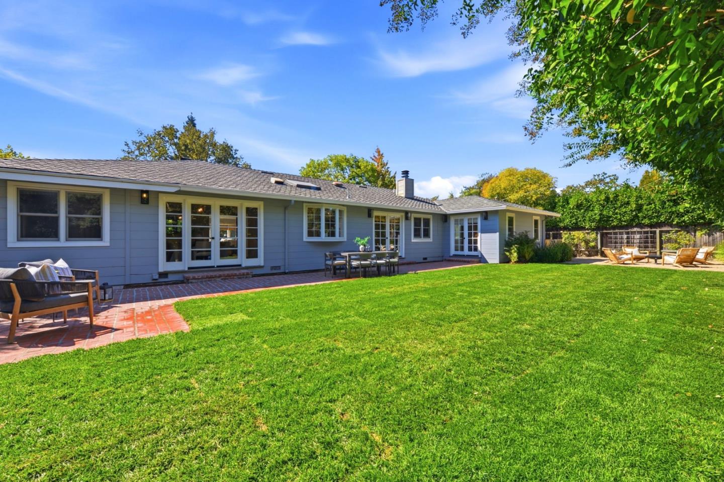Rear of a blue-gray single-story home with a brick patio, outdoor seating, and a broad green lawn.